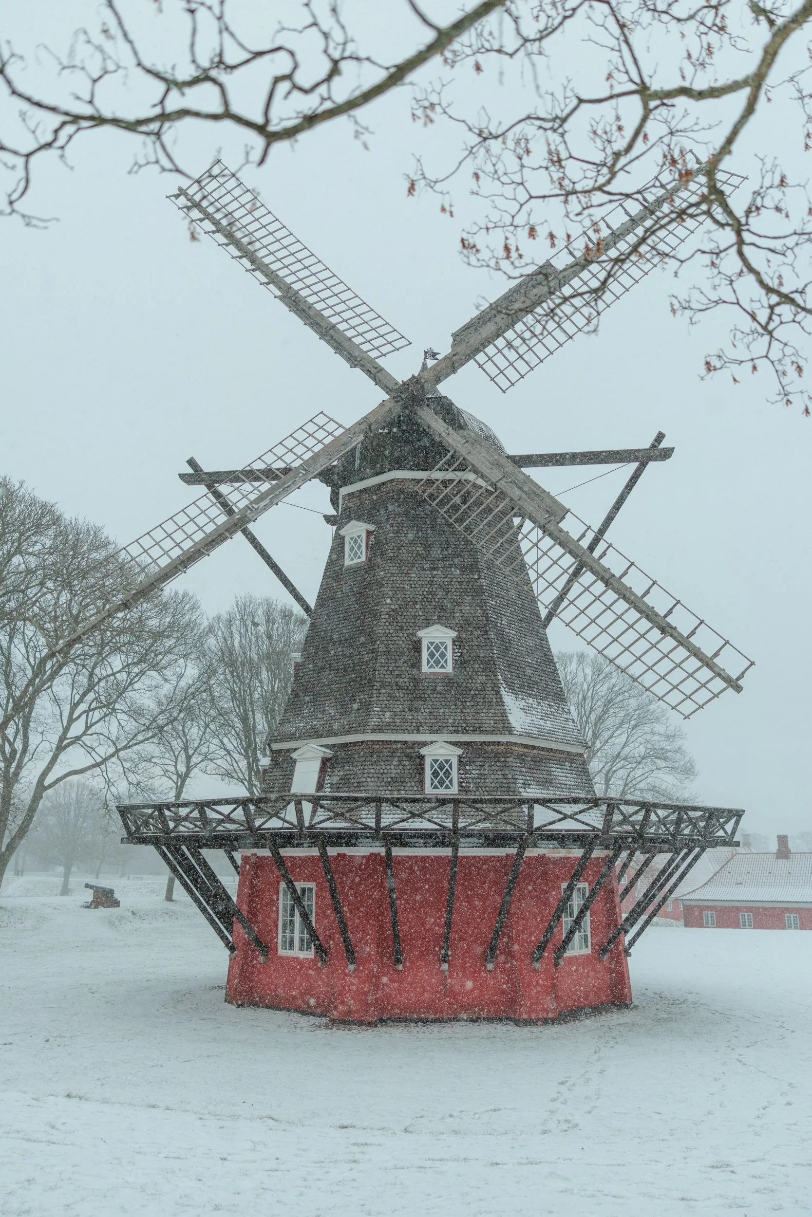 A traditional windmill with a wooden structure and red base standing in a snowy landscape, surrounded by leafless trees and falling snow.