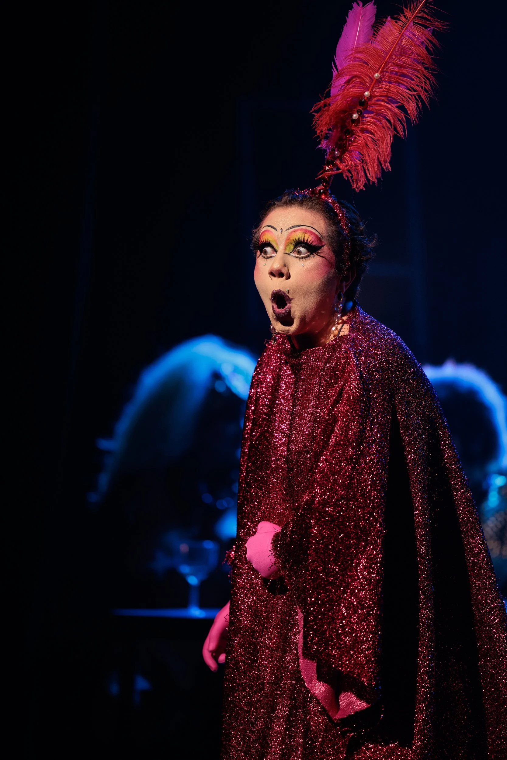 Performer with elaborate makeup, a large pink feathered hat with pearls, and a sparkly red gown on stage, with a dark background and blurred figure in the background.
