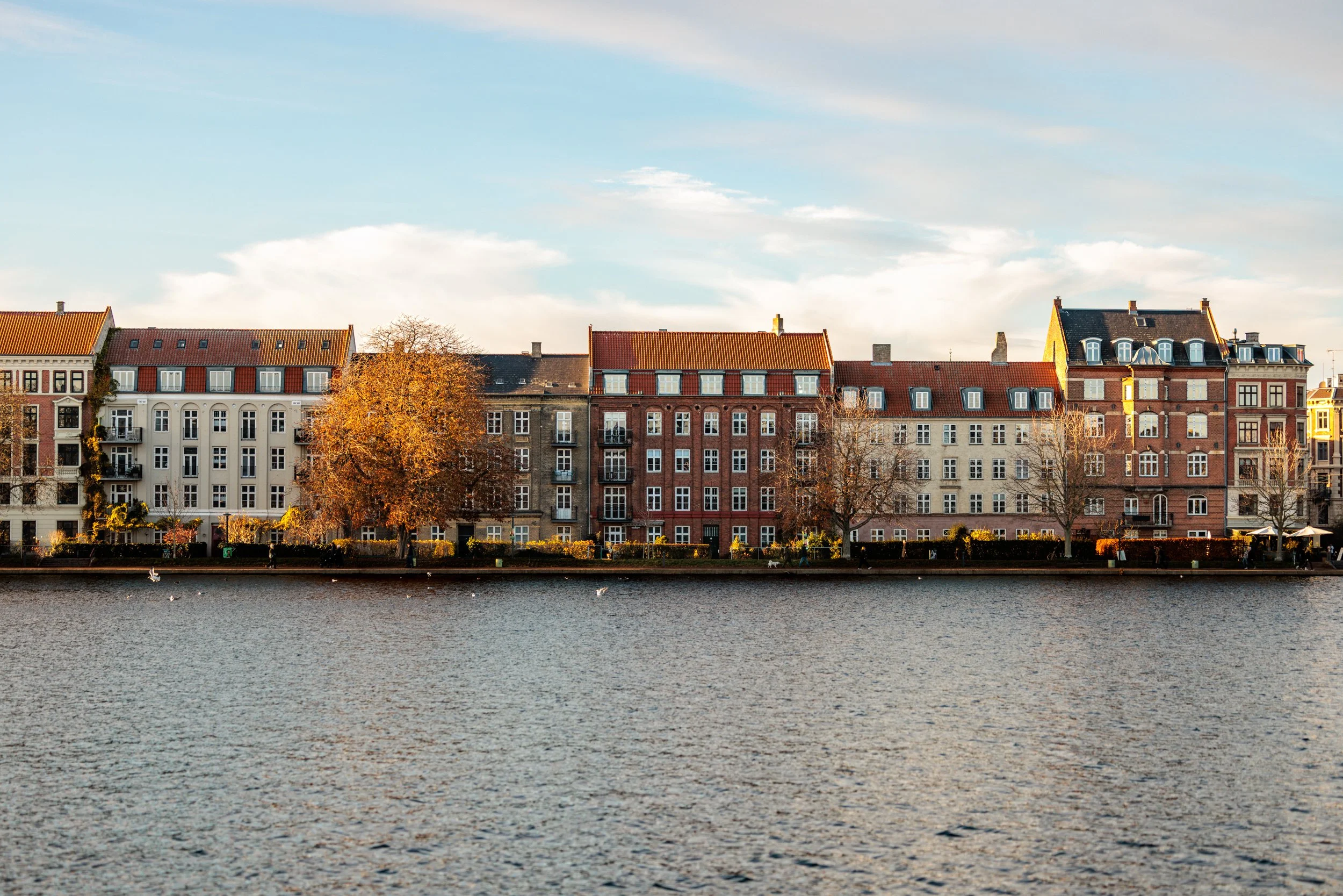 A row of colorful old European-style buildings with tiled roofs along a waterway, with trees in autumn foliage and a partly cloudy sky in the background.