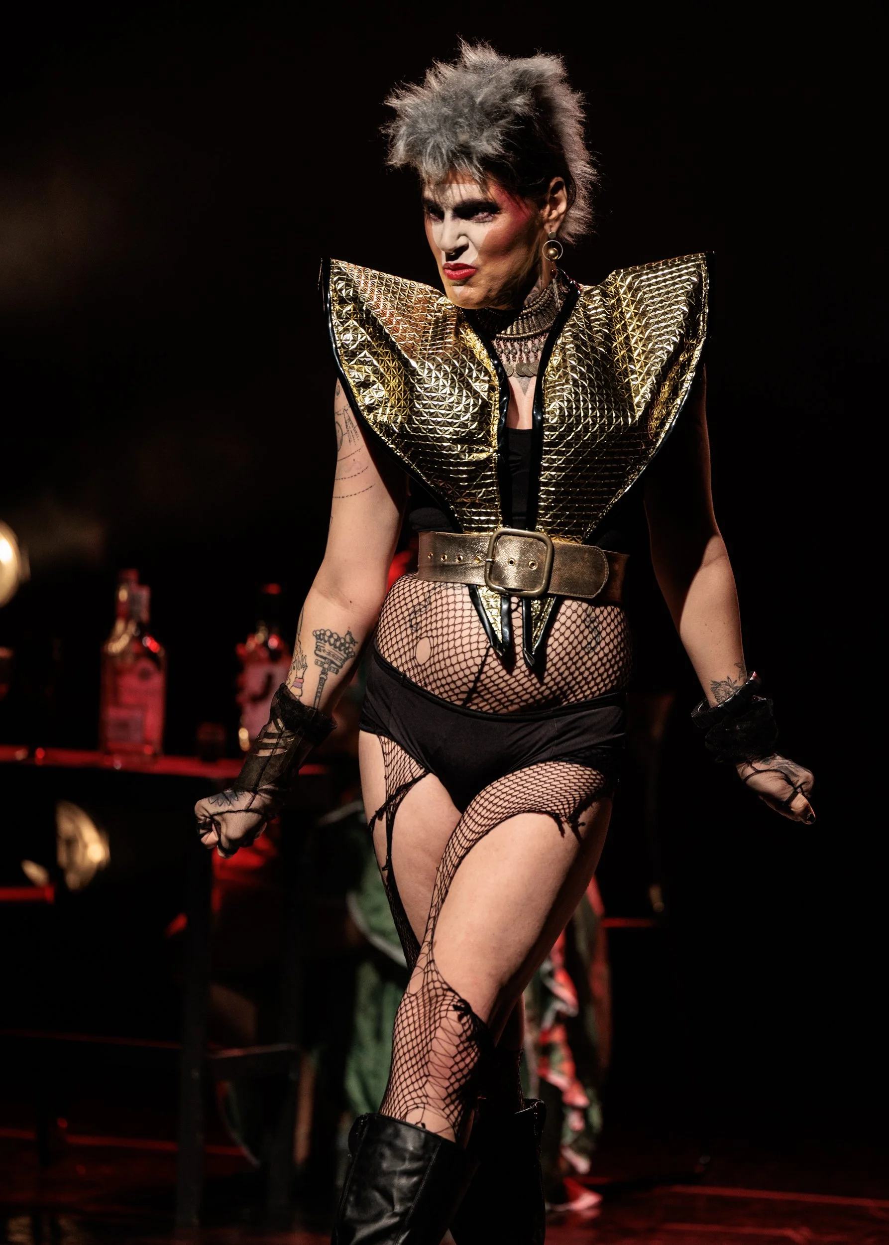 A performer on stage wearing a metallic gold geometric-patterned shoulder piece, fishnet stockings, black gloves, black shorts, and tall black boots, with short spiky gray hair and bold makeup.