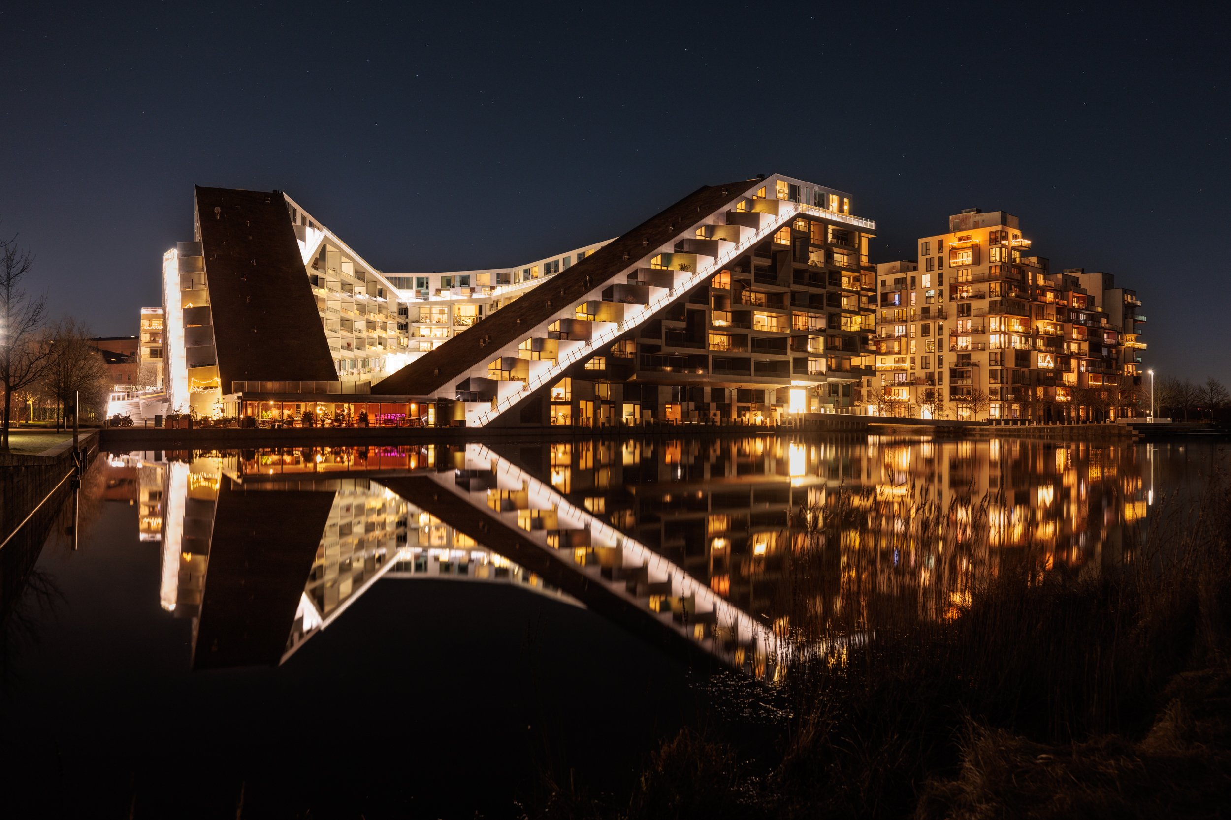 Night view of modern residential building with illuminated windows, reflected in a body of water in the foreground.