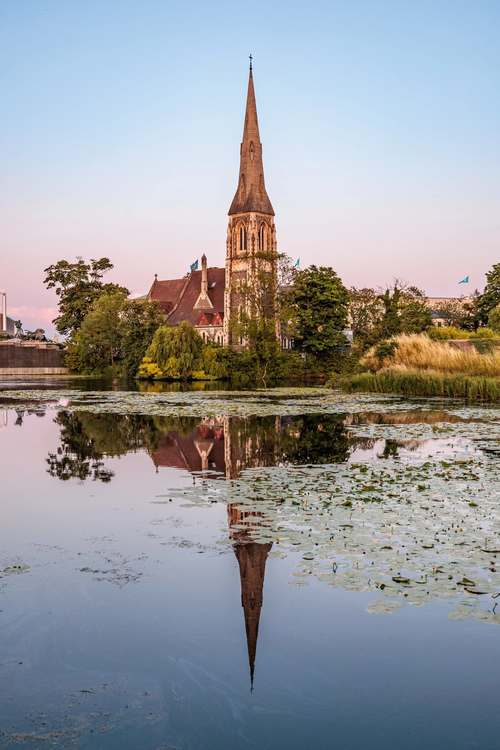 A church with a tall, pointed steeple reflected in the water surrounded by trees and lily pads.