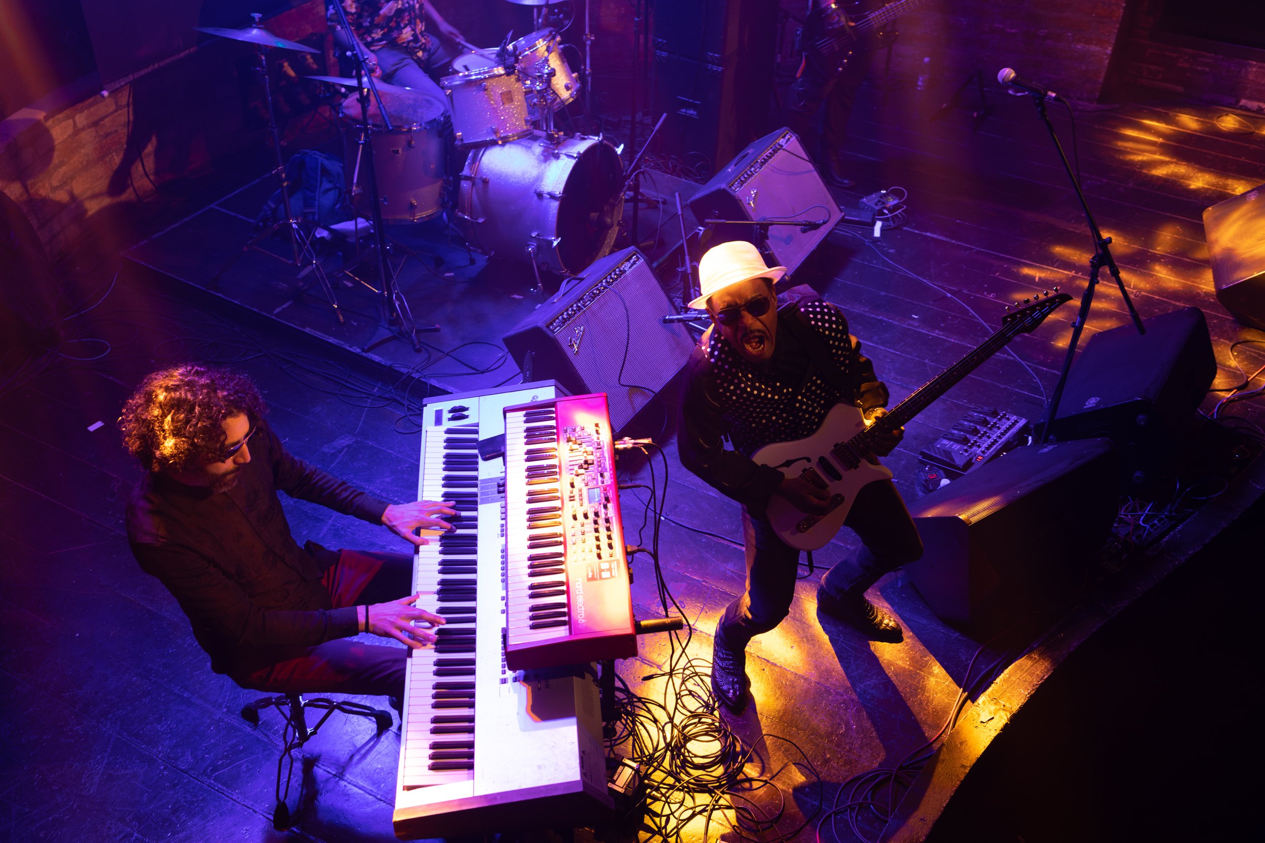 Live musical performance featuring a keyboardist, guitarist, and drummer on stage under stage lighting.