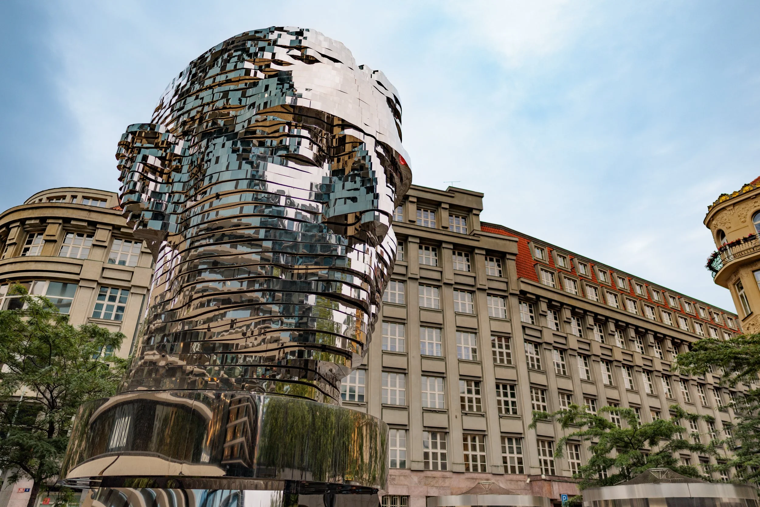 A large modern sculpture of a human face made of reflective, layered metal panels in an outdoor urban setting.