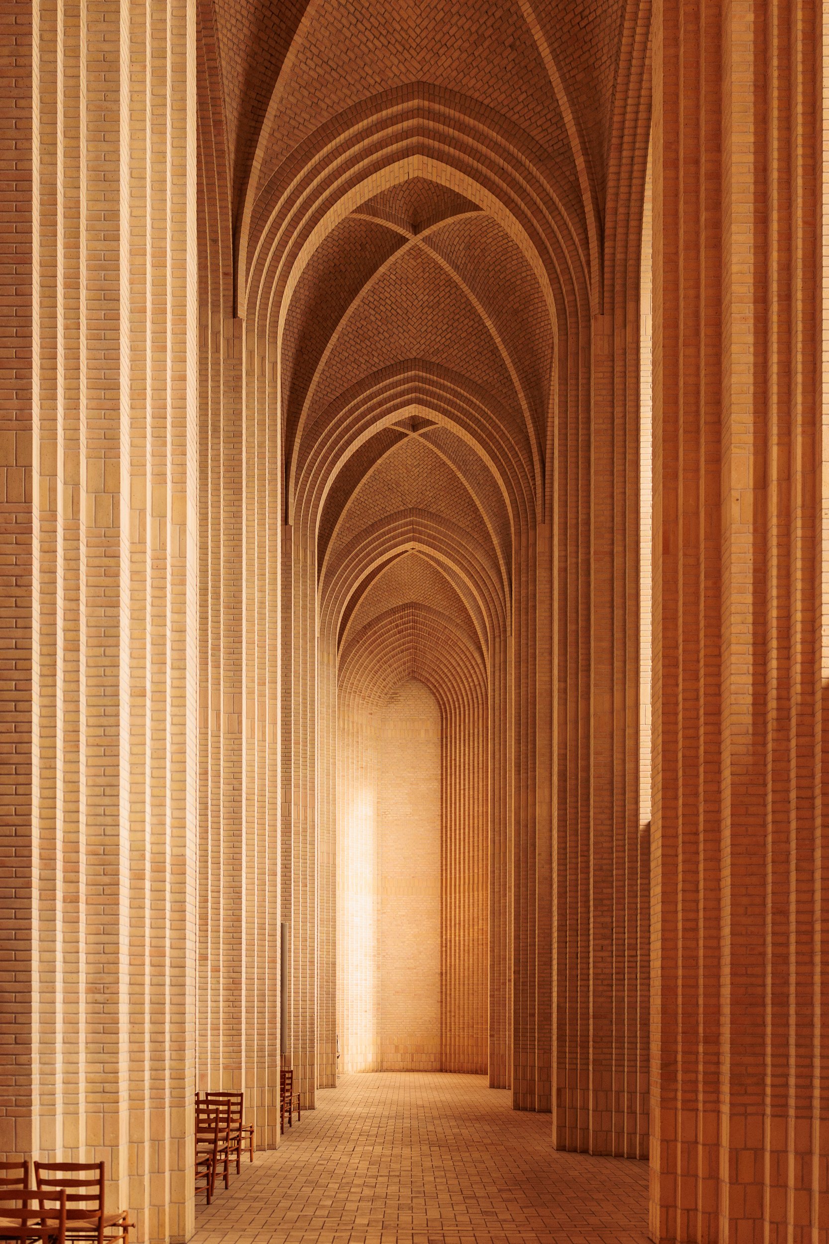Perspective view of a large brick corridor with high arched ceilings and columns on both sides, lit by warm lighting, with chairs along the left side.