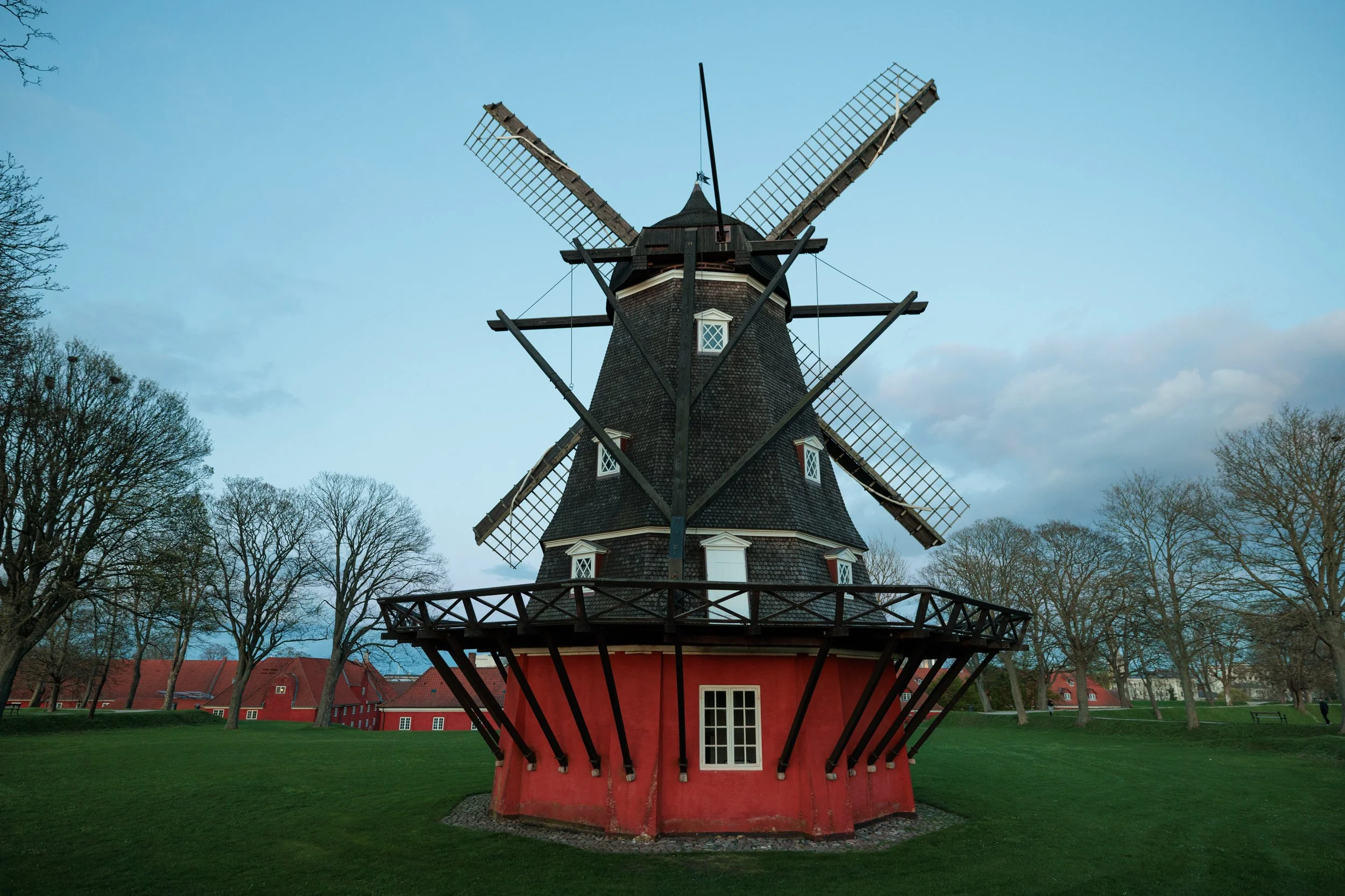 A traditional windmill with a red base and dark wooden upper structure surrounded by leafless trees and green grass under a blue sky.
