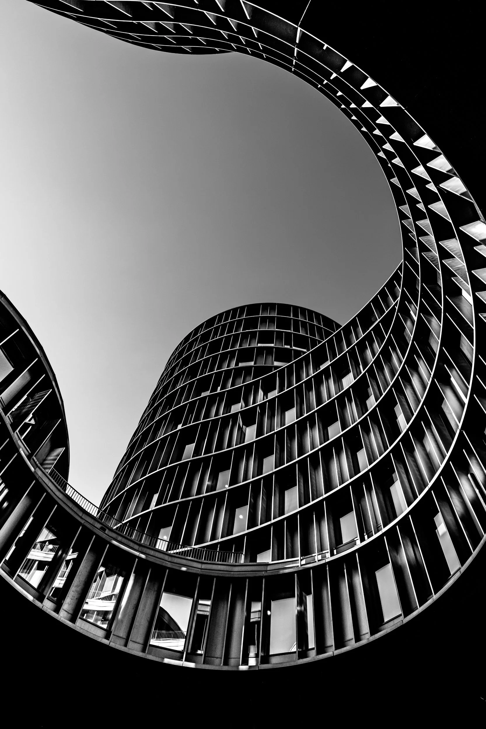 View of a modern, curved glass and metal building from the ground, looking up at the sky through the open center.