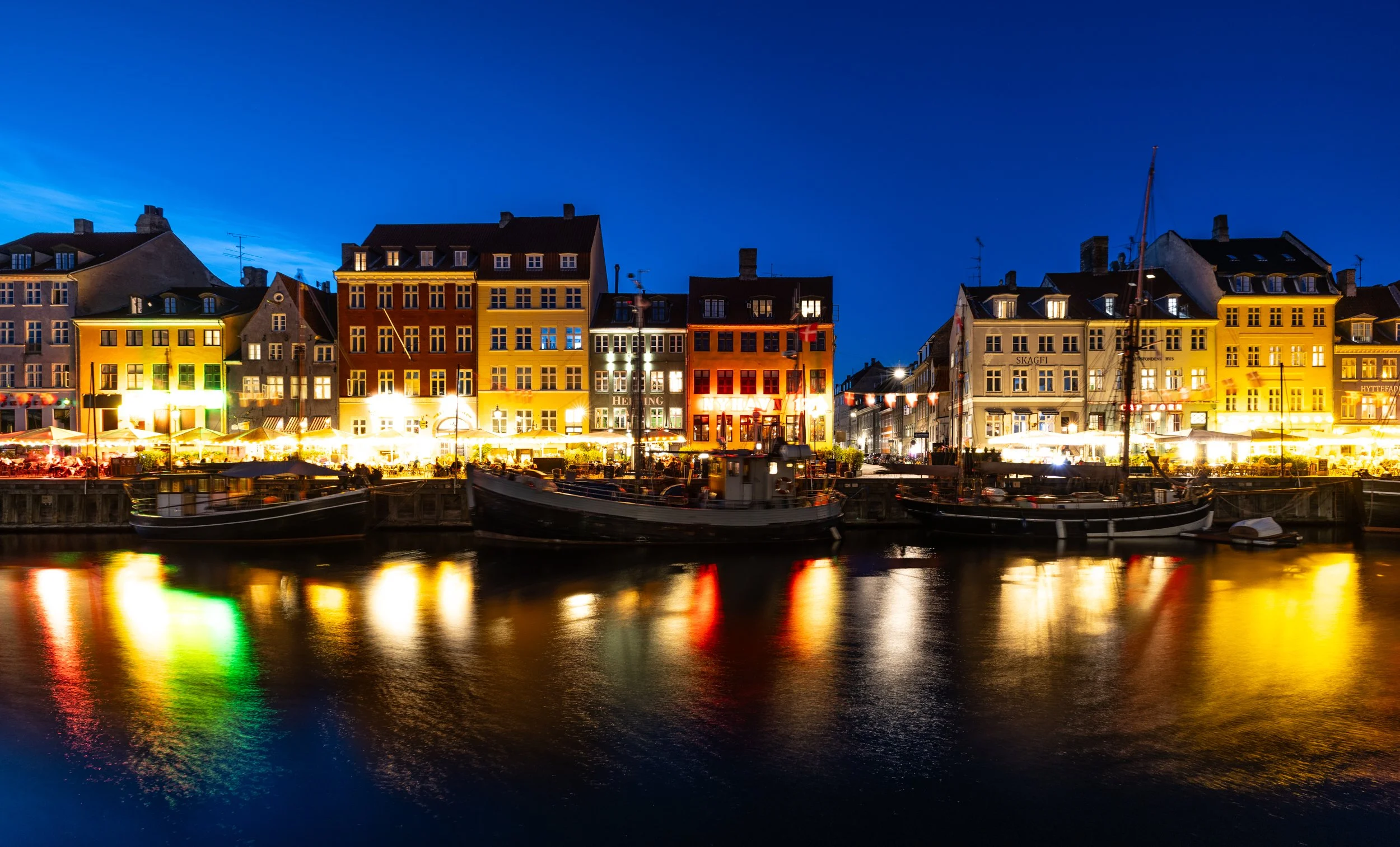 Colorful buildings with illuminated windows and outdoor lights along a waterfront dock, with boats reflecting on the water at night under a clear dark blue sky.