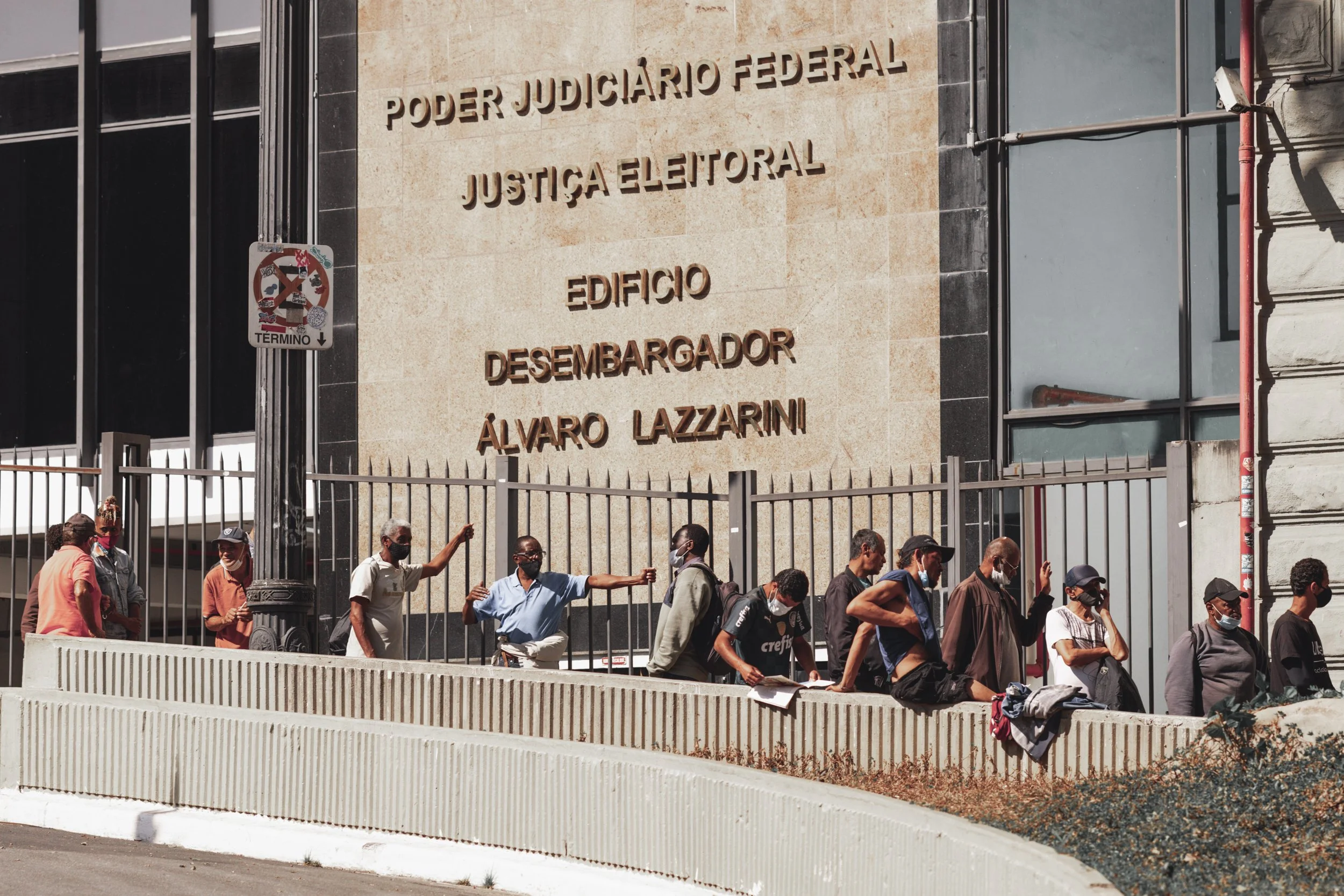 People walking past the entrance of a courthouse building with a sign that reads "Poder Judicial Federal Justiça Eleitoral Edificio Desembargador Álvaro Lazzarini." Some individuals are wearing masks, and a few are engaging in conversations as they w