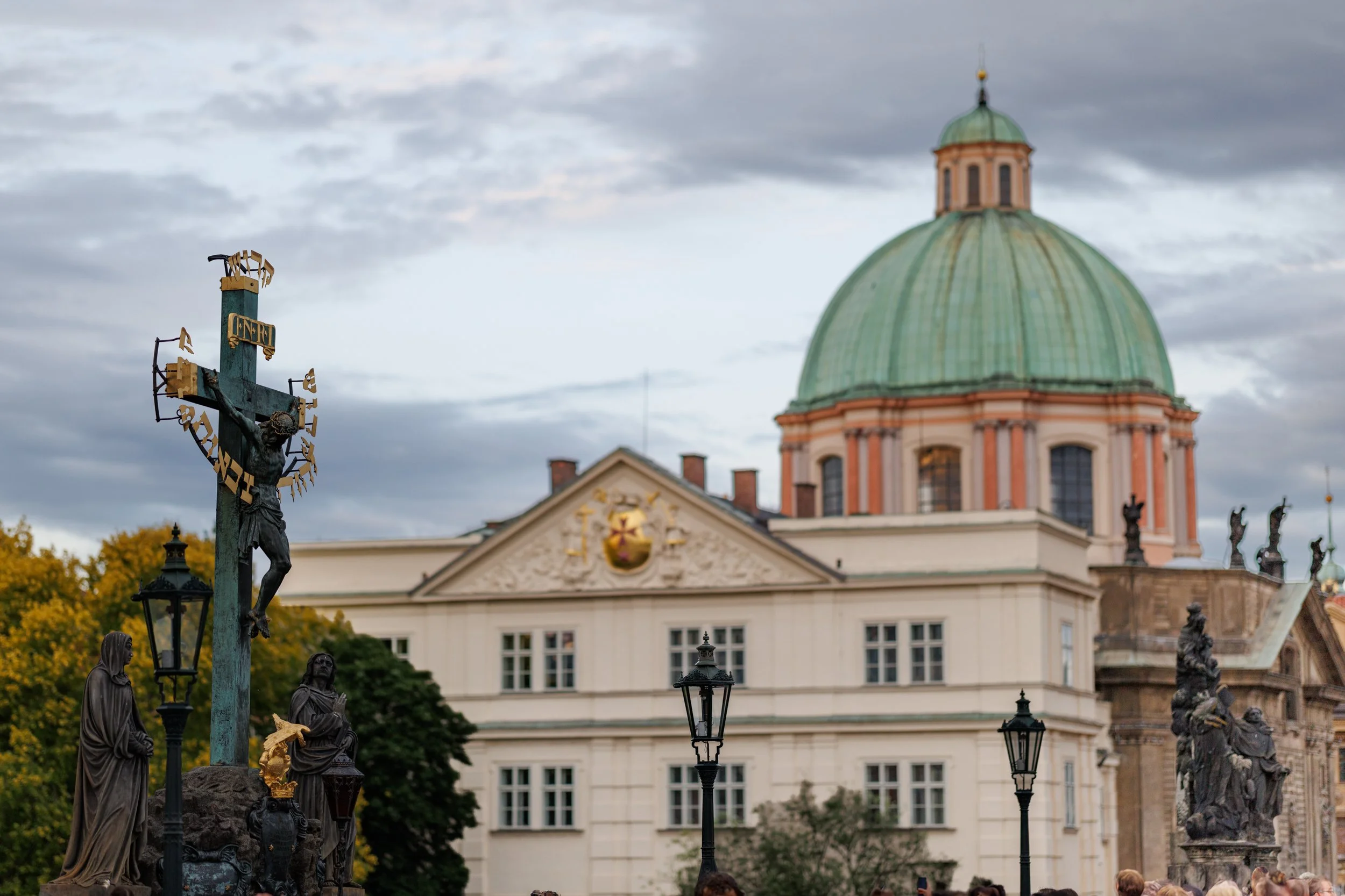 Statues depicting religious figures and a crucifix at a historic site, with a classical building having a large green dome in the background under a cloudy sky.