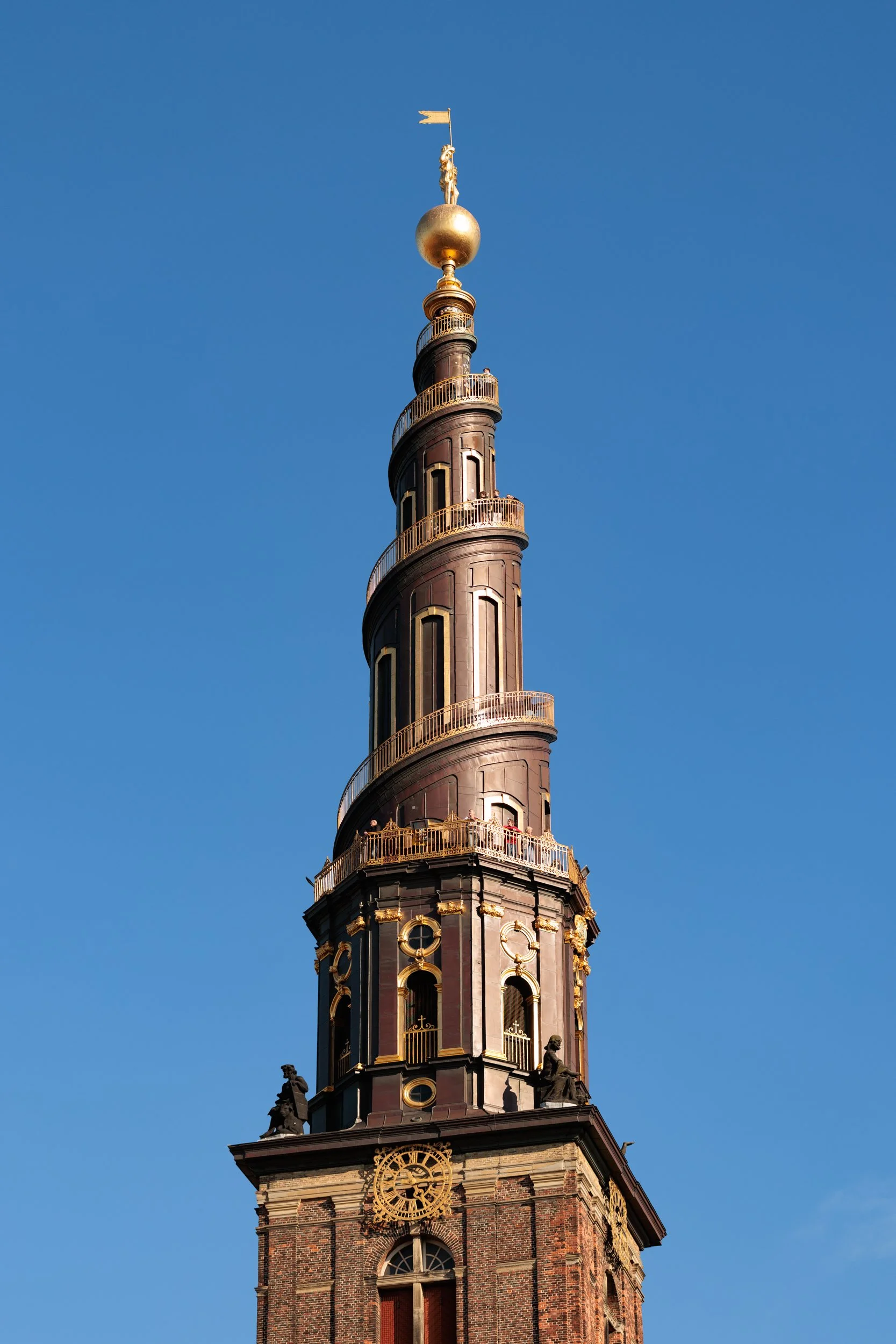 A historic clock tower with a dark brick facade, golden decorative accents, and a tall spire topped with a gold sphere and statue, set against a clear blue sky.