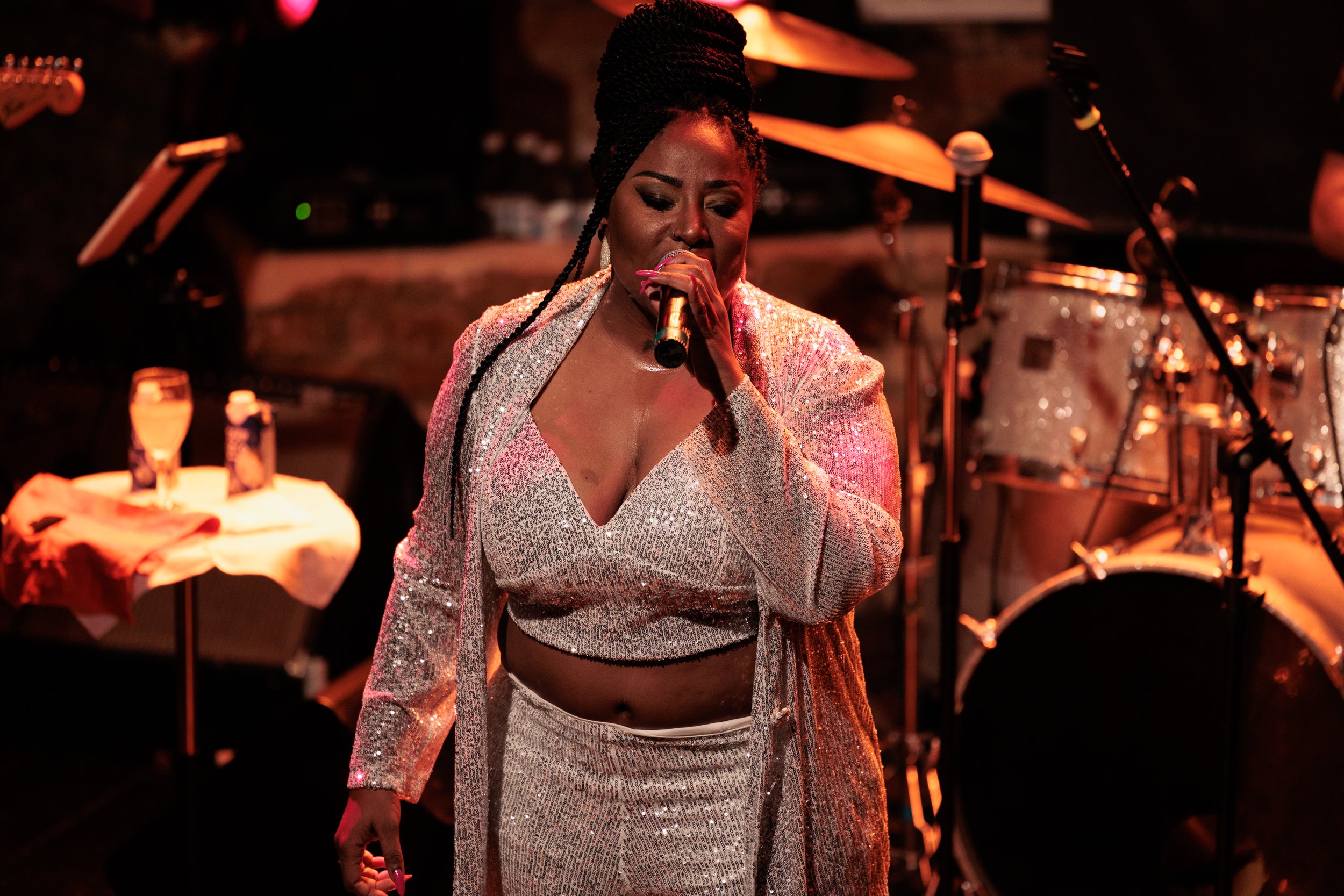 A woman singing into a microphone at a music event. She has dark braided hair styled in an updo and is wearing a shiny, silver, sequined outfit with a matching skirt, exposing her midriff. The background includes musical instruments like drums and mi