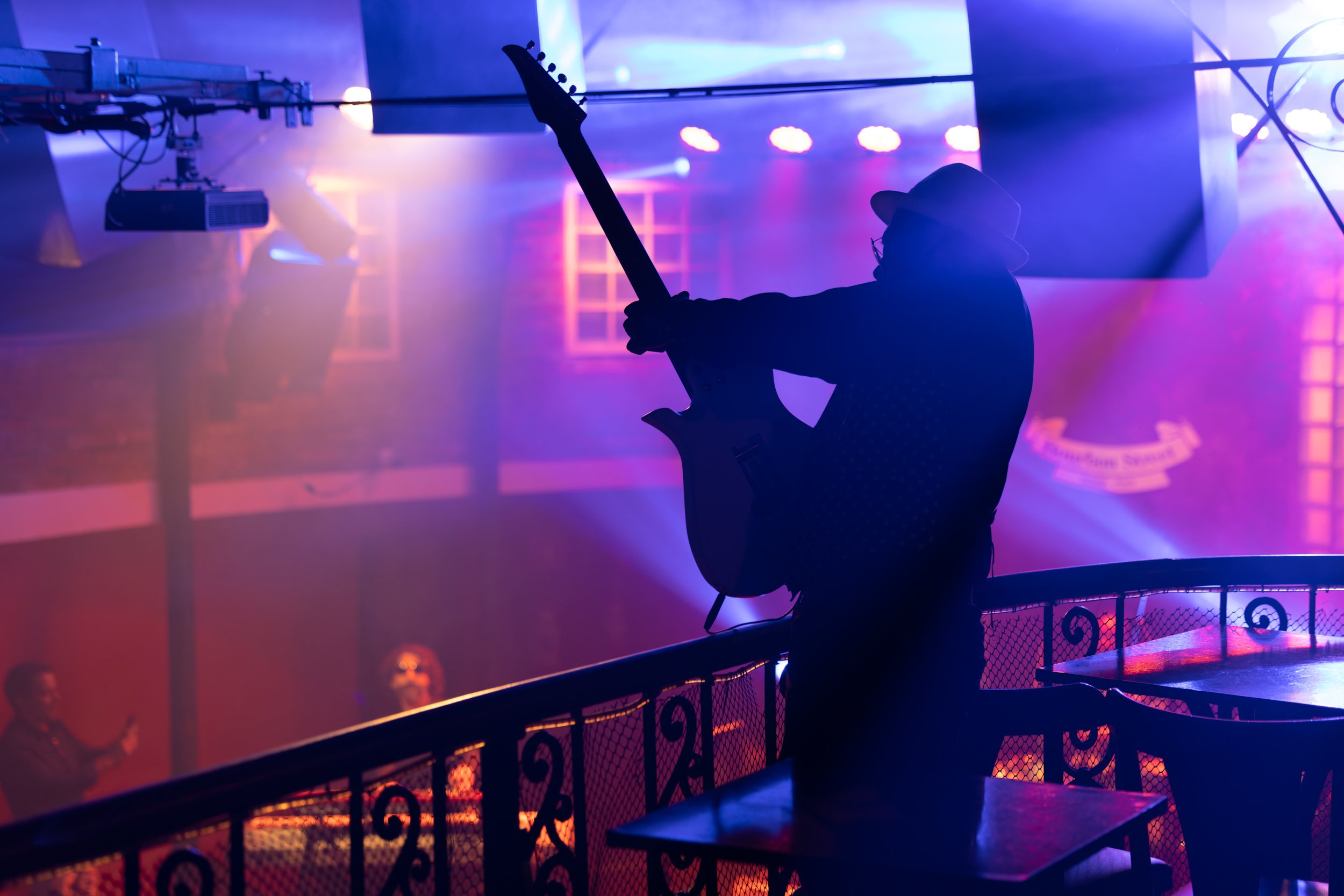 Silhouette of a person wearing a hat and glasses, playing an electric guitar on stage with colorful blue and purple stage lights, in a concert or nightclub setting.