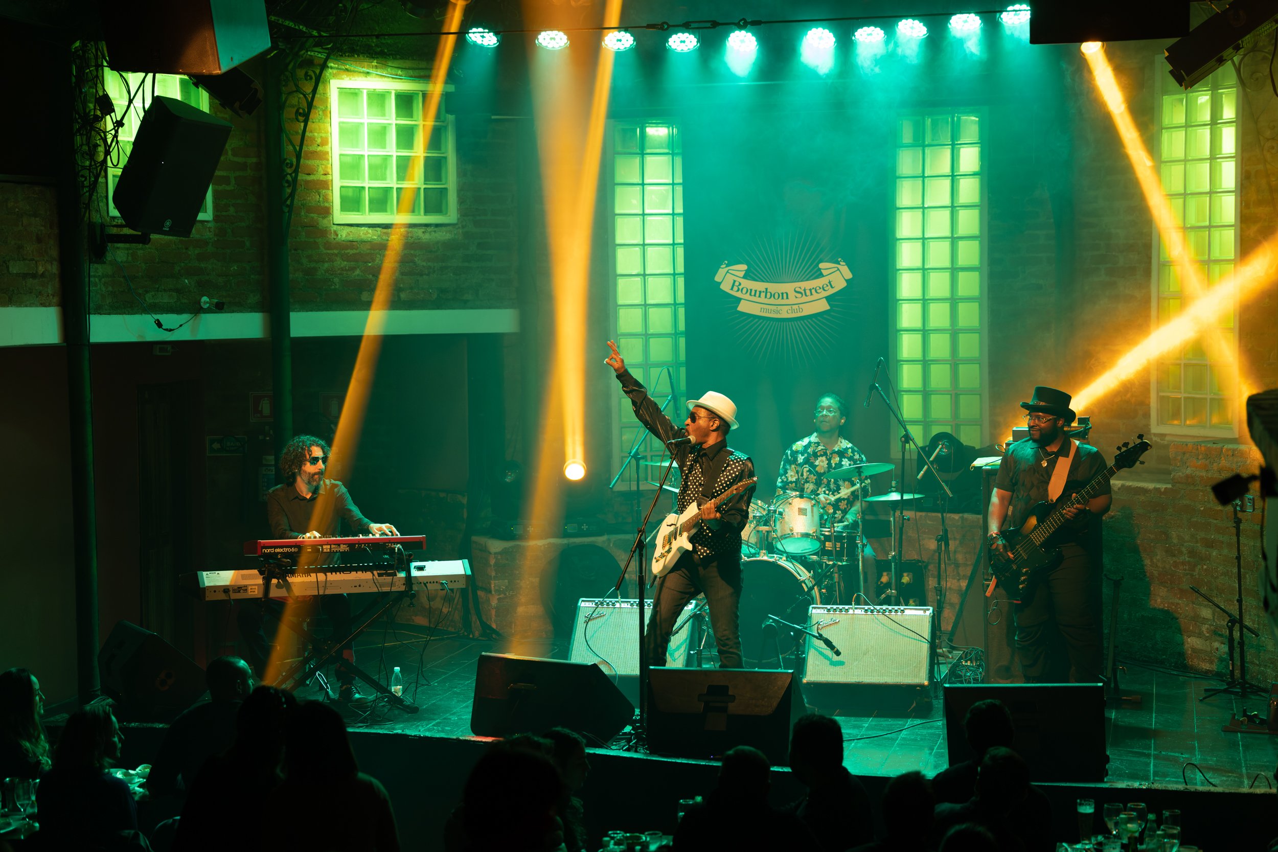 A band performing on stage at Bourbon Street music club with green and yellow stage lighting and brick walls, with an audience seated in front.