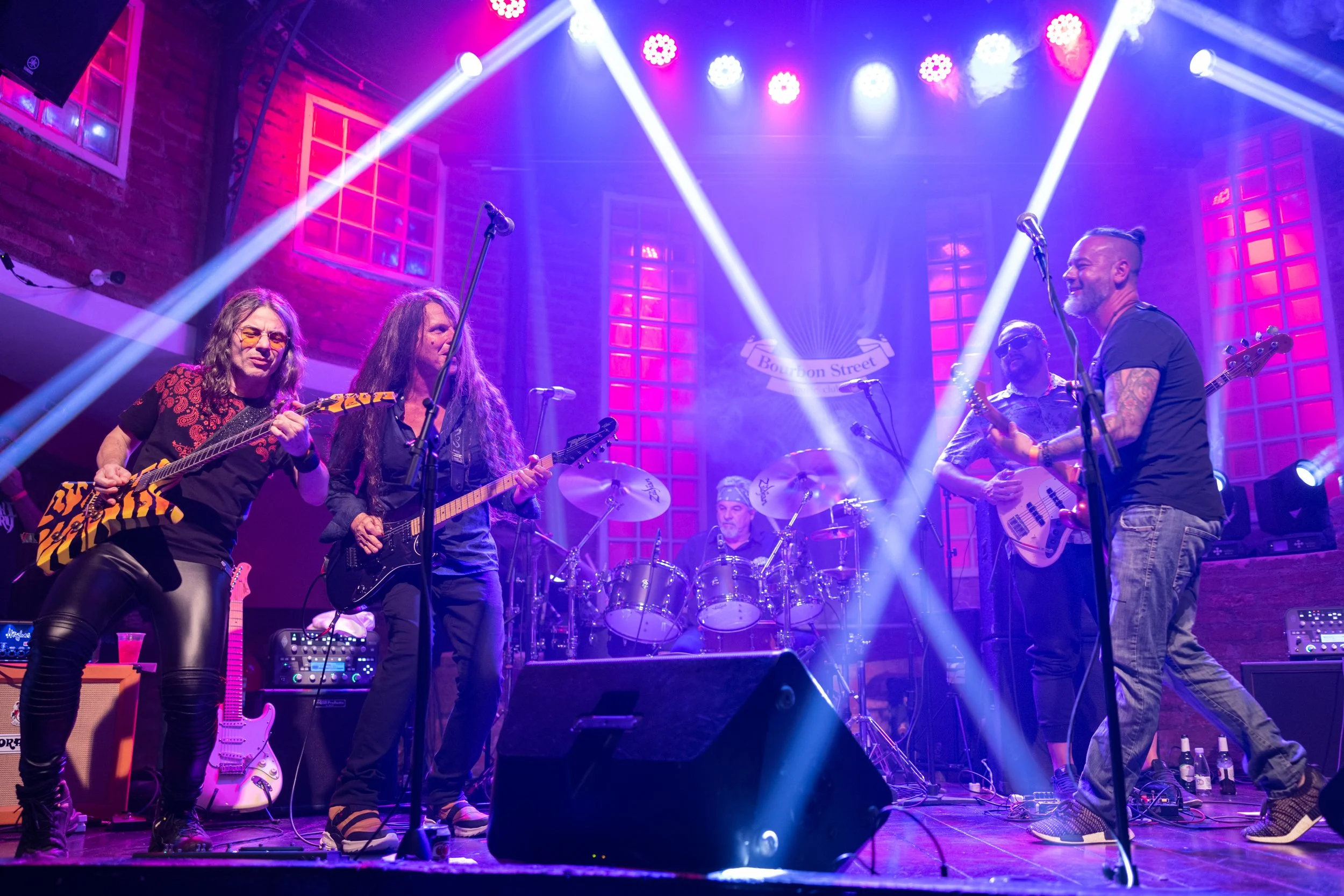 A live music performance with five band members on stage, playing guitars and drums, illuminated by colorful stage lights in a cozy venue with brick walls and windows.