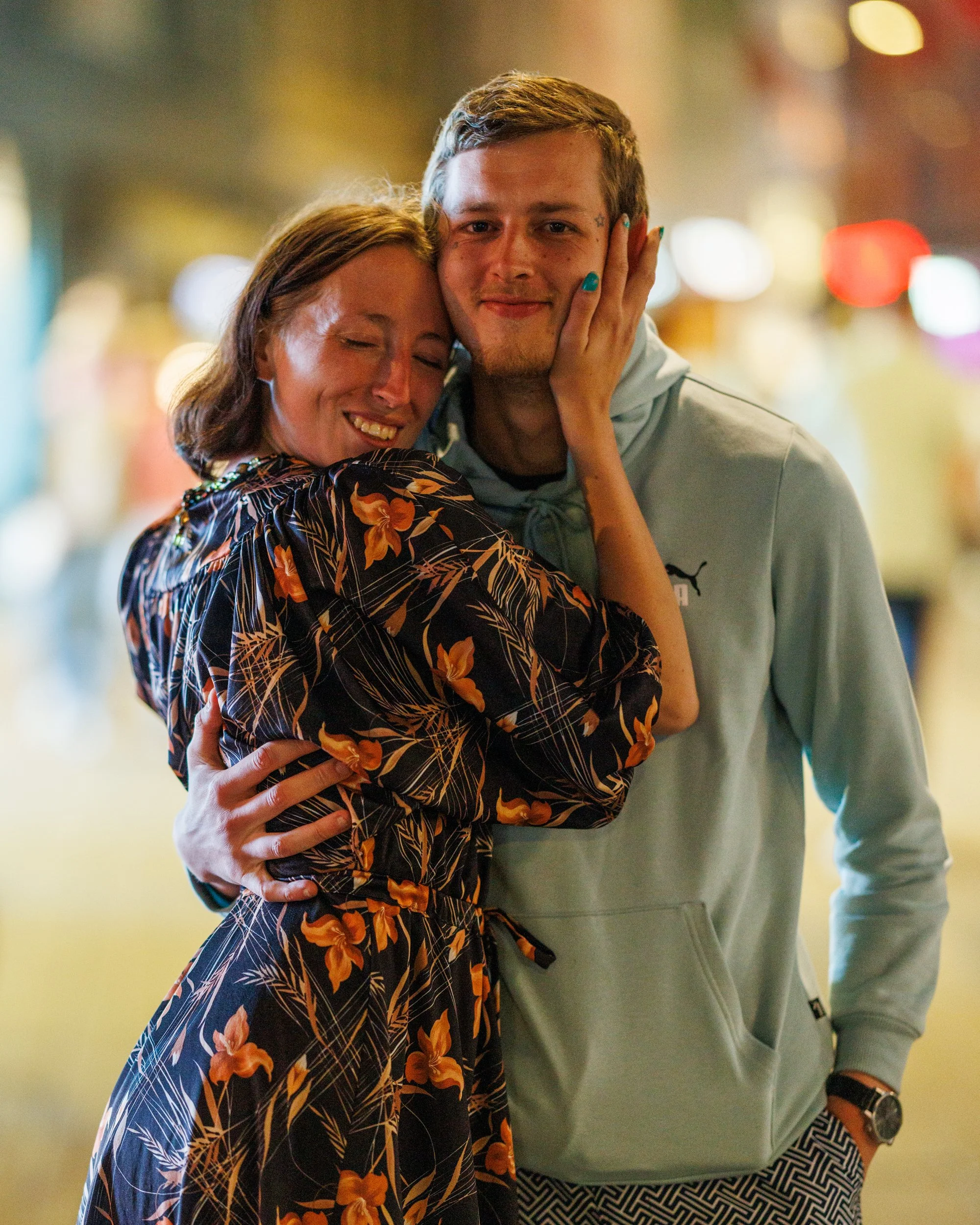 A woman with shoulder-length brown hair embracing a man with short light brown hair. The woman is smiling with eyes closed, and the man is smiling directly at the camera. They are holding each other's faces affectionately in a blurred indoor setting 