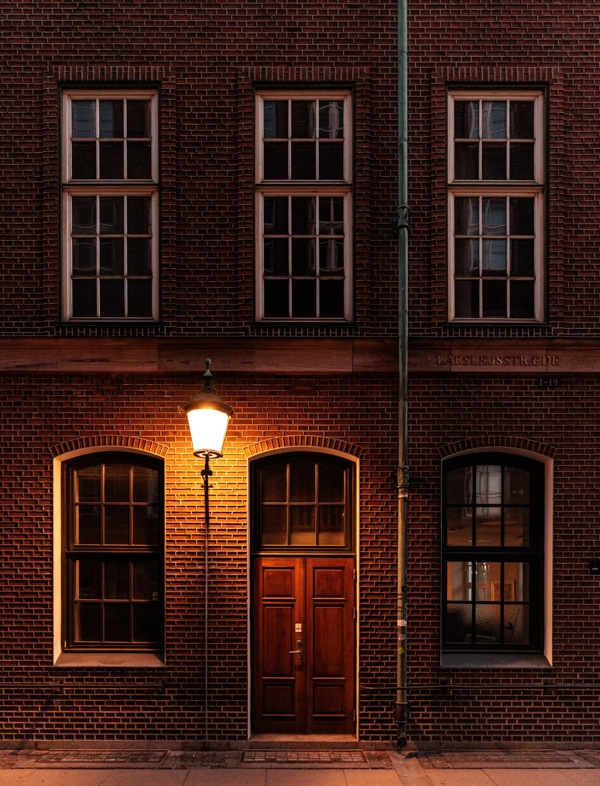 A brick building with three tall windows on the second floor, a large wooden door on the ground floor, and a street lamp illuminating the sidewalk in front. There is a vertical pipe running in front of the building and the street sign reads "Larslejs