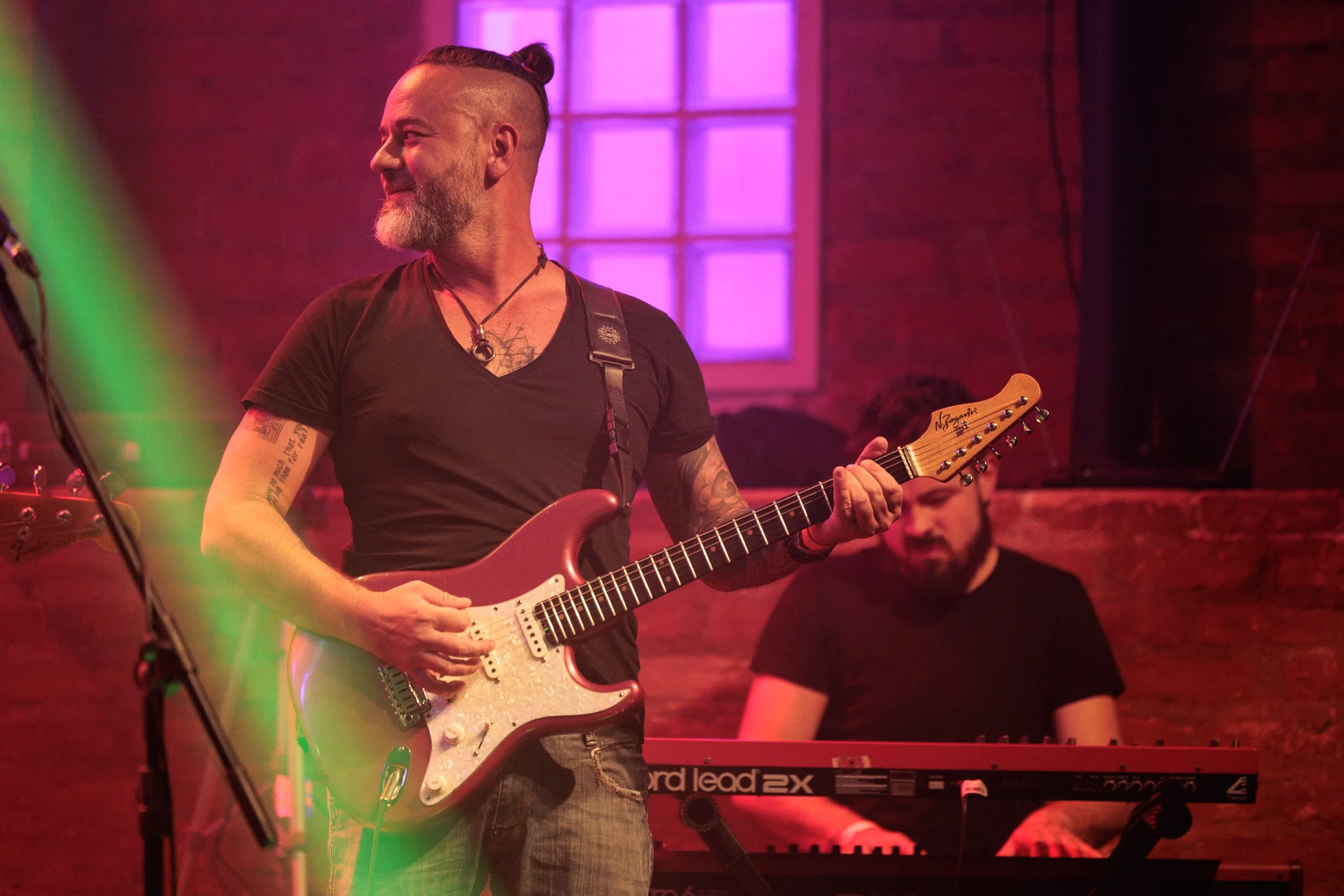 A man with a beard and tattoos playing an electric guitar on stage. There is a keyboard in front of him and a colorful, neon-lit background.