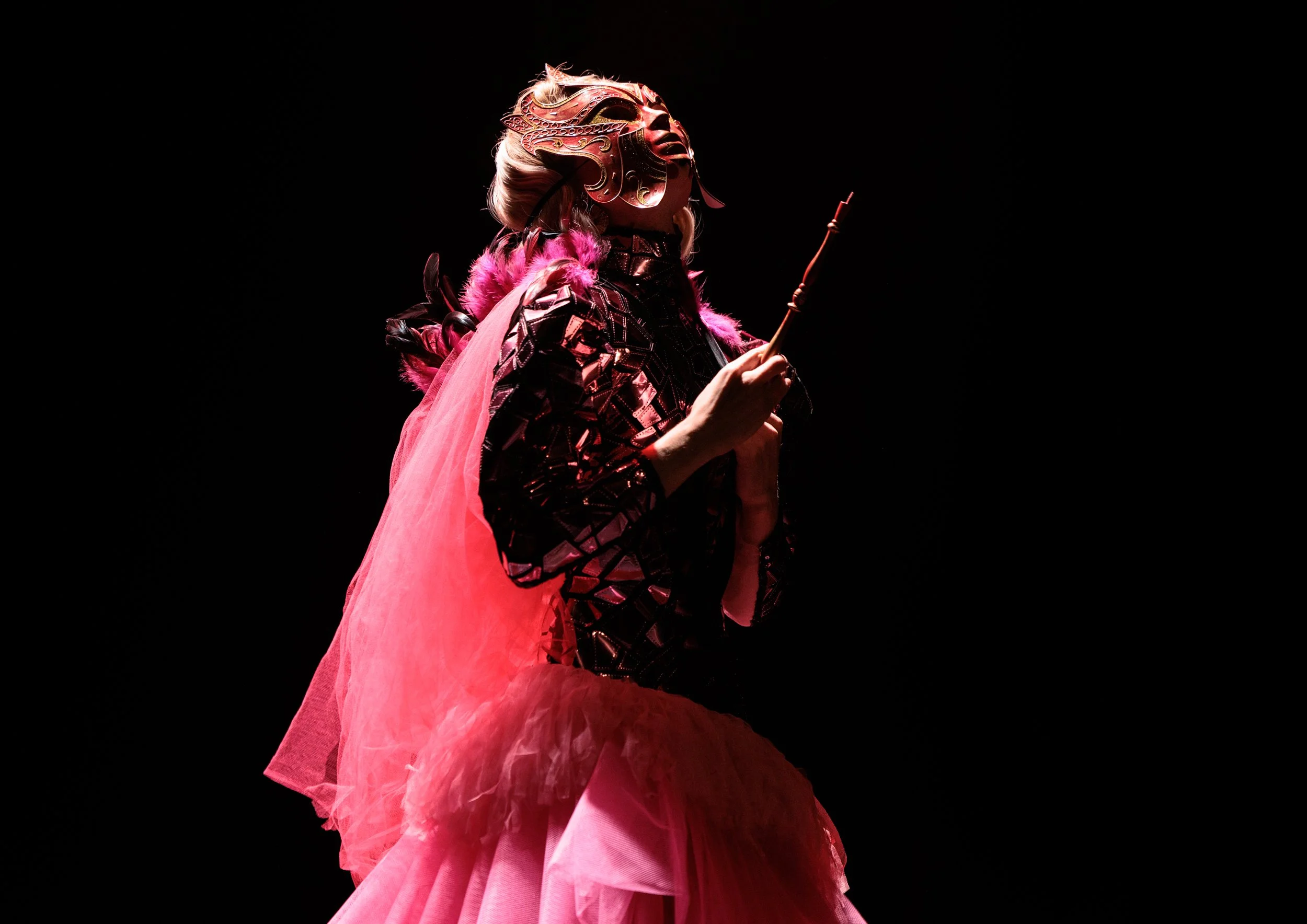 A person in a pink gown with a flowing skirt, wearing a metallic mask with intricate designs and a crown-like headpiece. They hold a wand and are set against a dark background, illuminated by a spotlight.