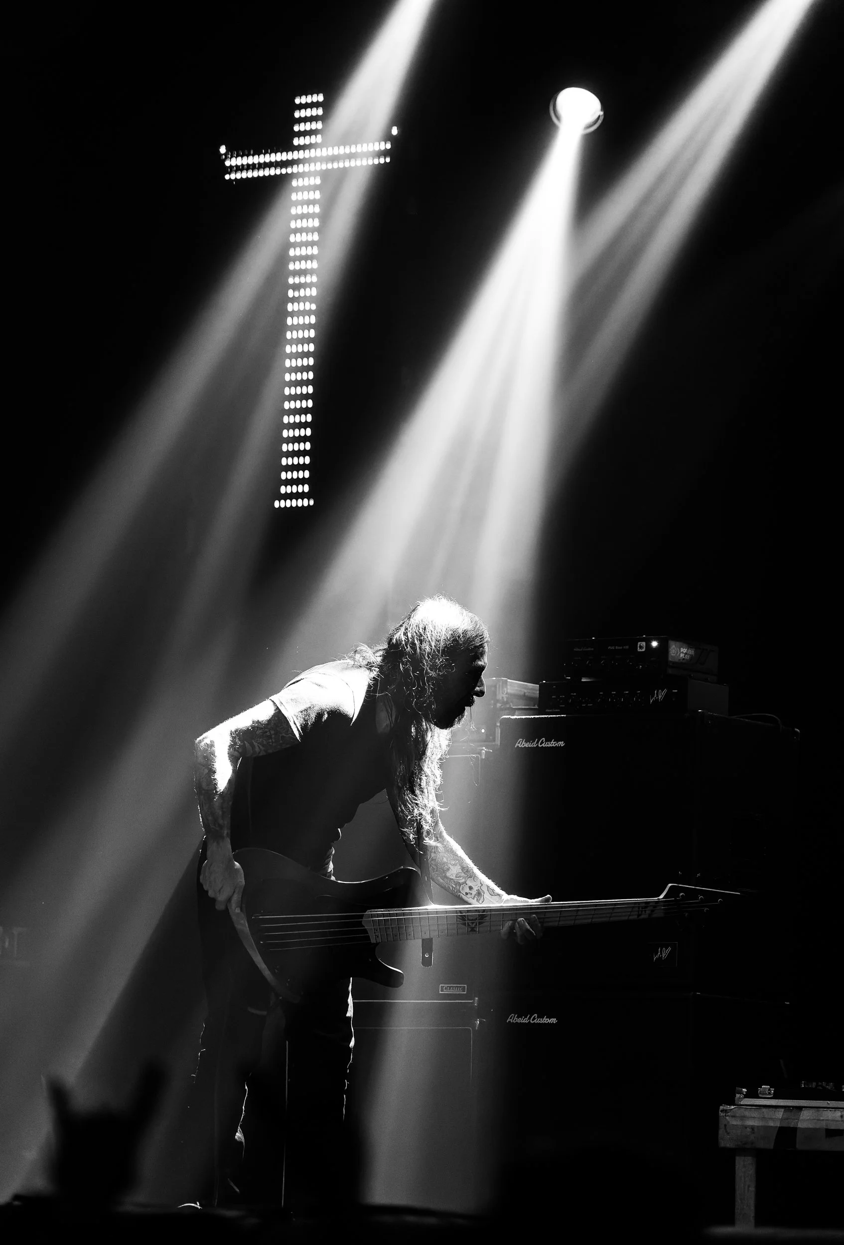 A musician with long hair and tattoos bending over an electric guitar on stage. Bright spotlights shine down, creating a dramatic black and white scene.