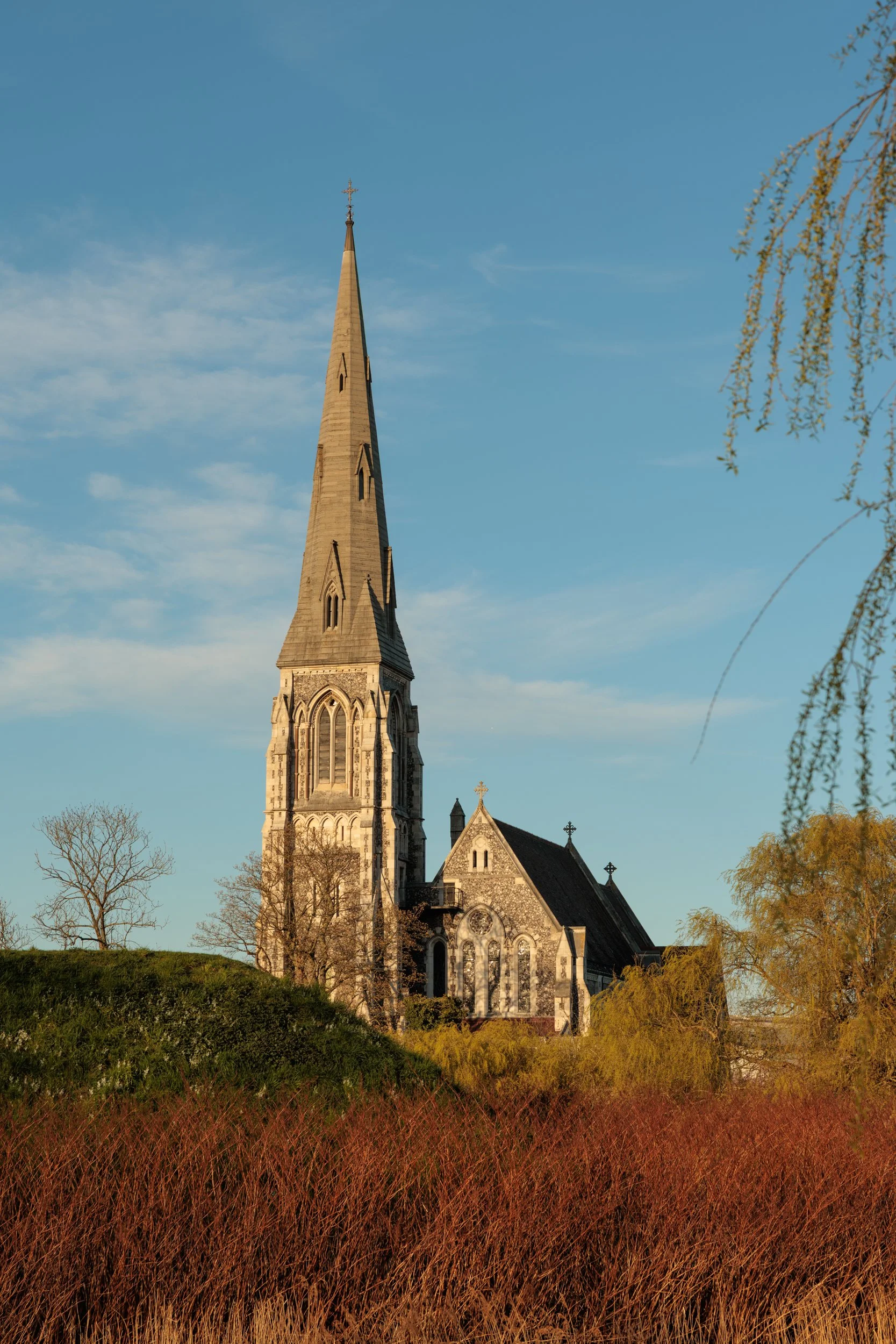 A church with a tall steeple and Gothic architectural features, surrounded by trees and grassy landscape, under a clear blue sky.