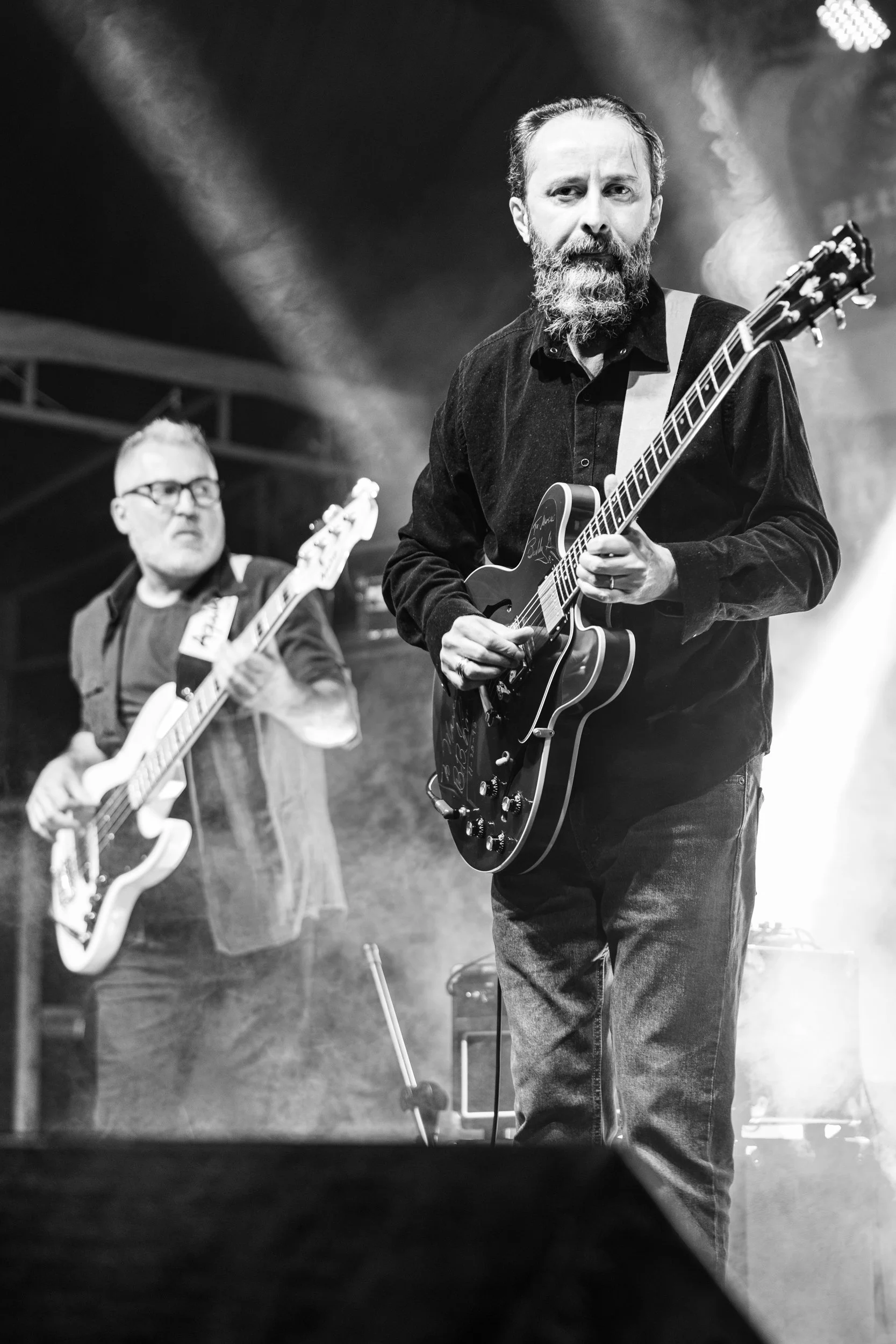 Black and white photo of two musicians playing guitars on stage, with stage lighting and haze. The man in the foreground has a beard, mustache, and is wearing a dark shirt, focusing on his guitar. The man in the background has glasses, a beard, and i