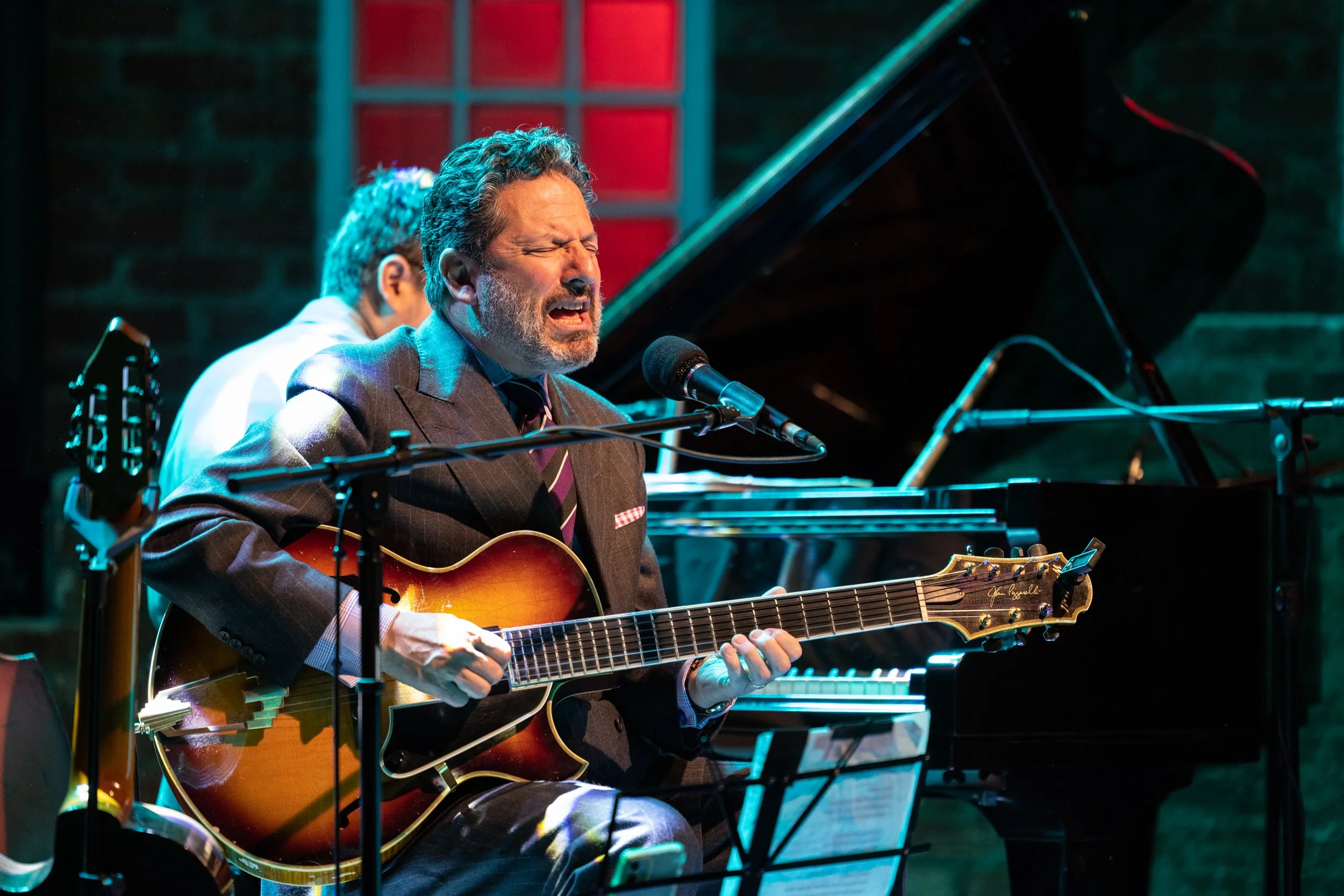 A man singing passionately while playing an acoustic guitar during a live performance, with a pianist visible in the background playing a grand piano.