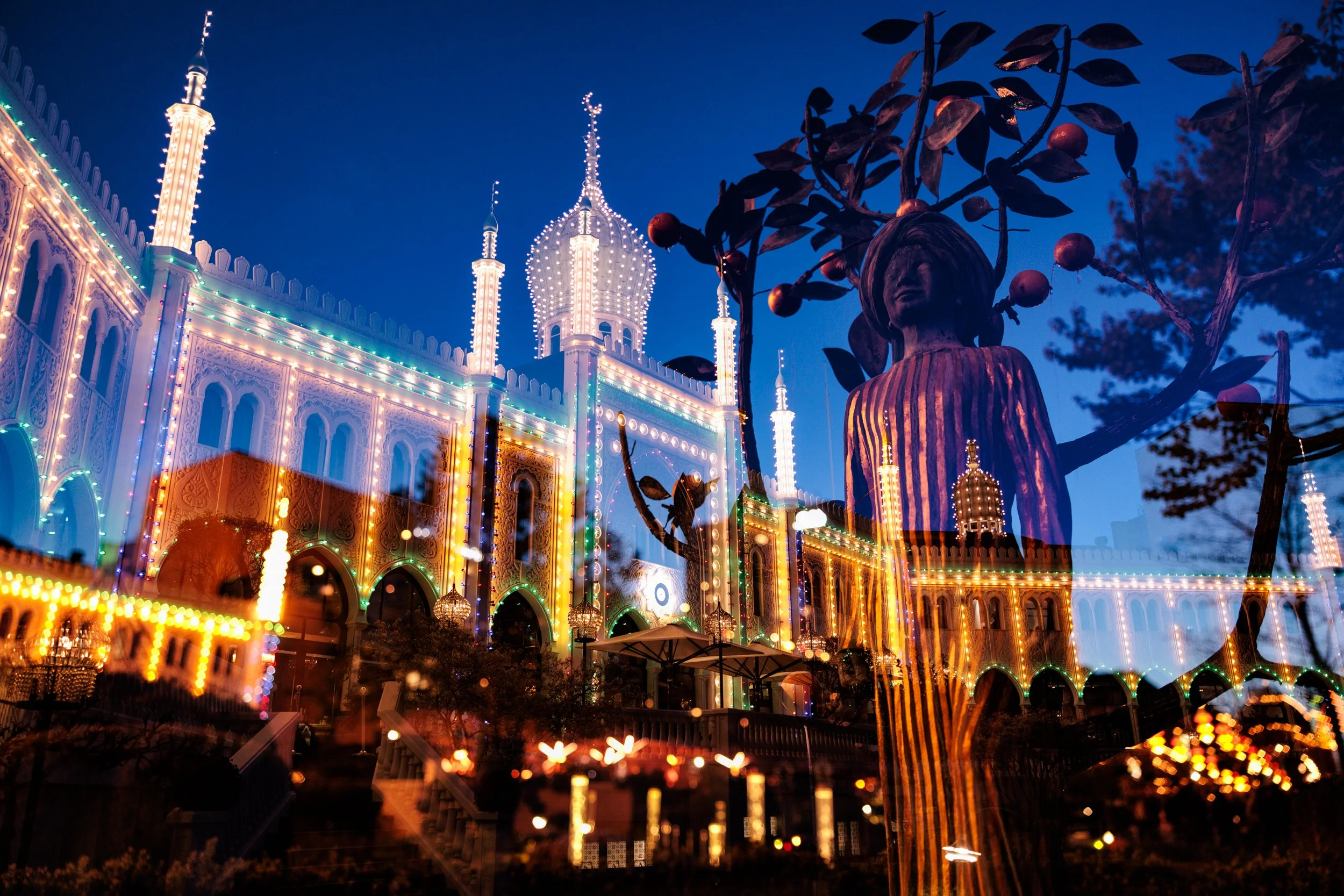 A brightly lit amusement park at night featuring a large ornate castle with colorful lights and multiple spires, with a statue of a girl with a fruit tree in the foreground.
