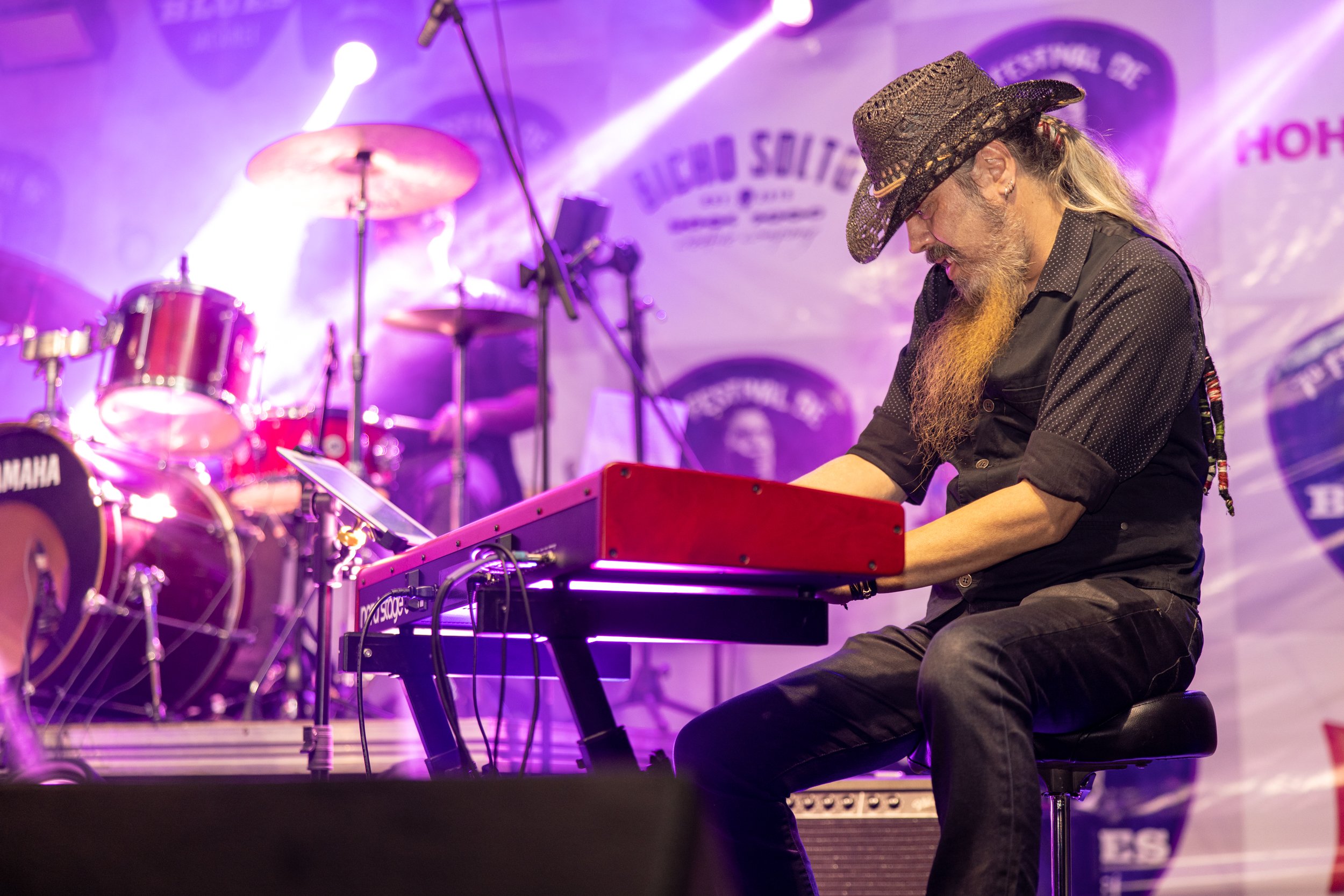 A man with a long beard wearing a cowboy hat and glasses playing a red keyboard on stage with purple lighting and a drum set in the background.