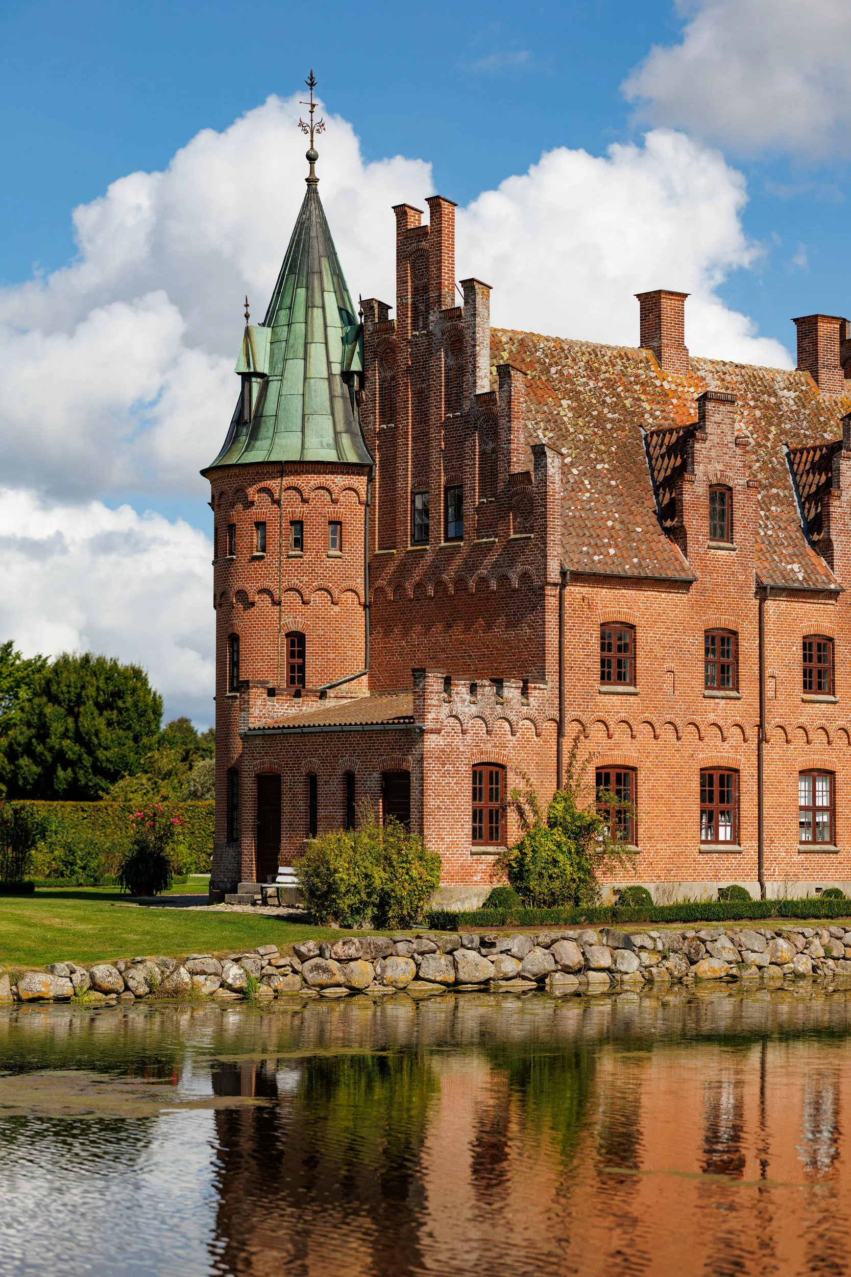 A historic red brick castle with a green copper spire, surrounded by a stone wall and greenery, reflecting in a body of water.