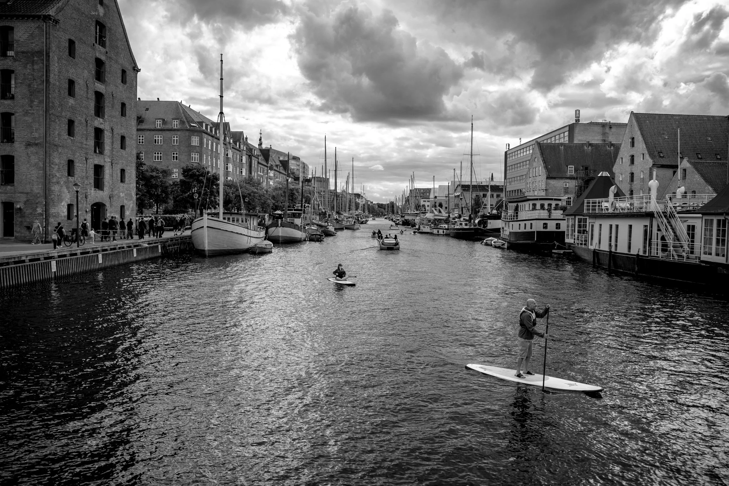 Black and white photo of a harbor with sailboats and buildings along the water, cloudy sky overhead, a person paddleboarding, and another person paddleboarding or standing on a paddleboard in the foreground.