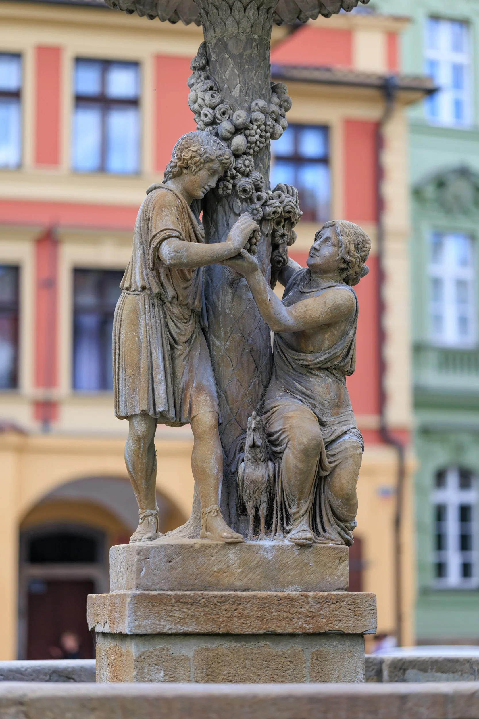 Close-up of a stone sculpture of two children playing, surrounded by a tree with carved fruits, set against a background of colorful buildings with large windows.
