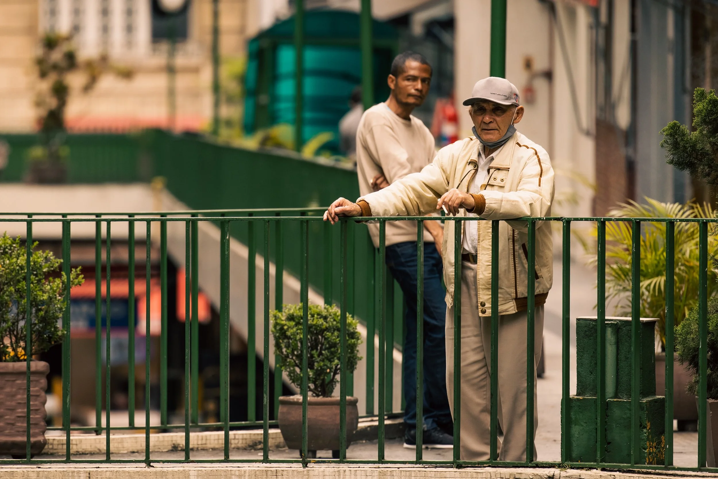 Two men standing behind a green metal railing on a city sidewalk, with potted plants in the foreground. One wears a beige jacket and a cap, the other wears a beige shirt.