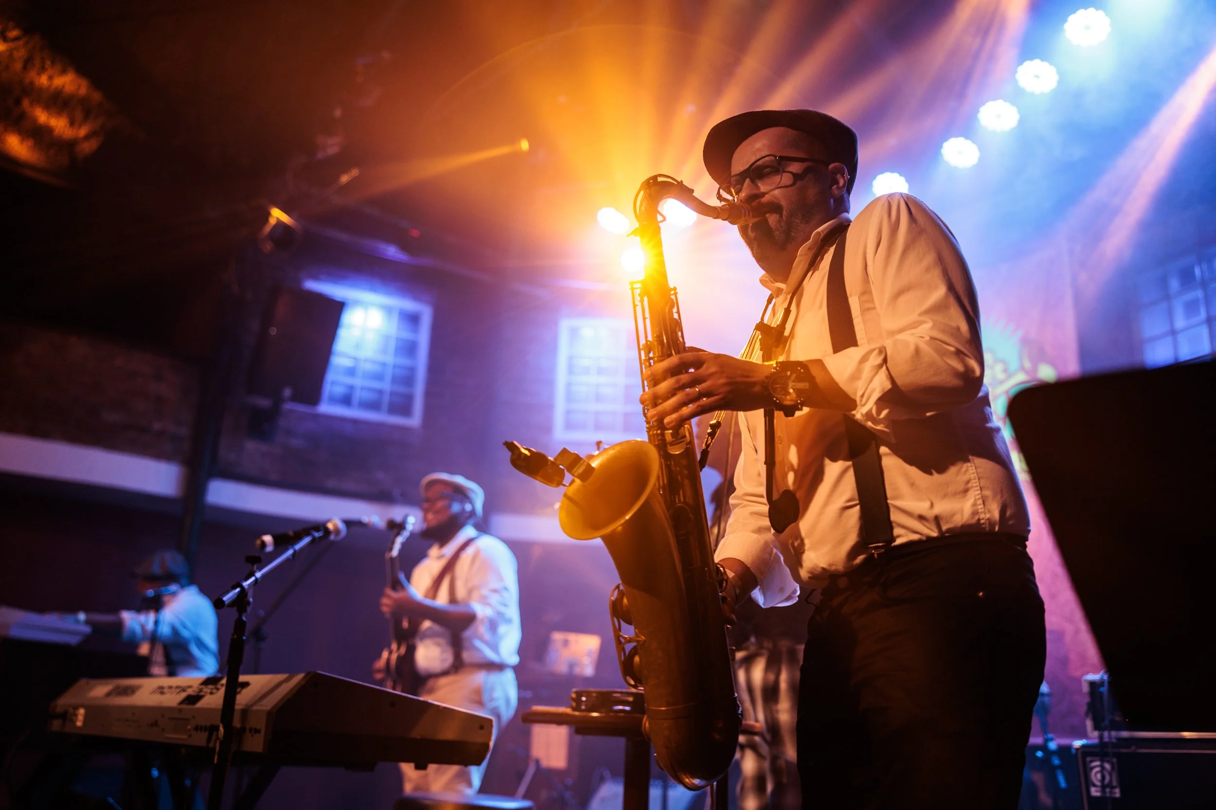 Musician playing saxophone on stage with colorful lighting, other band members playing keyboards and drums in the background.