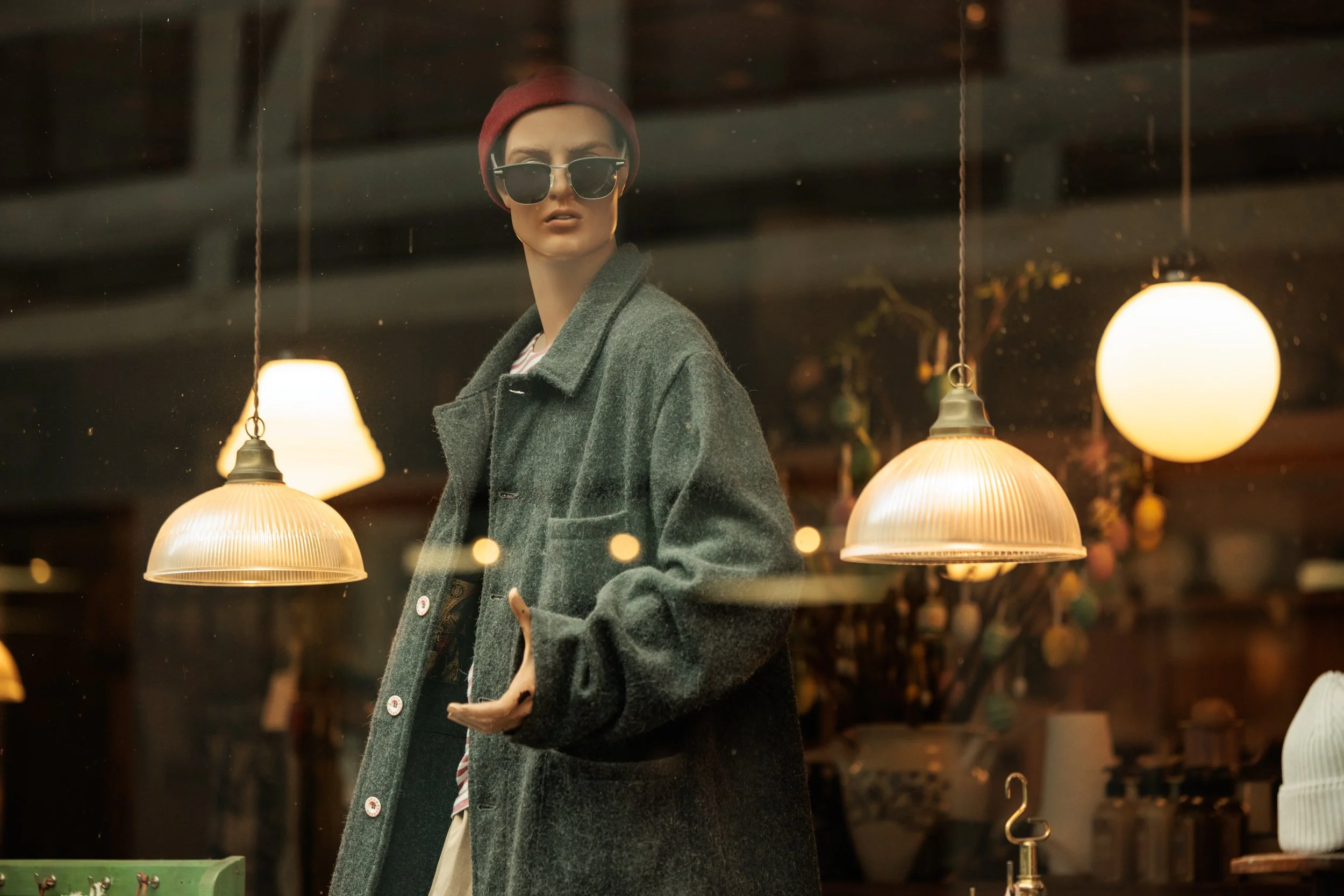 Mannequin with dark sunglasses, red beanie, gray coat, and striped shirt, displayed in a store window with hanging lamps and blurred background.