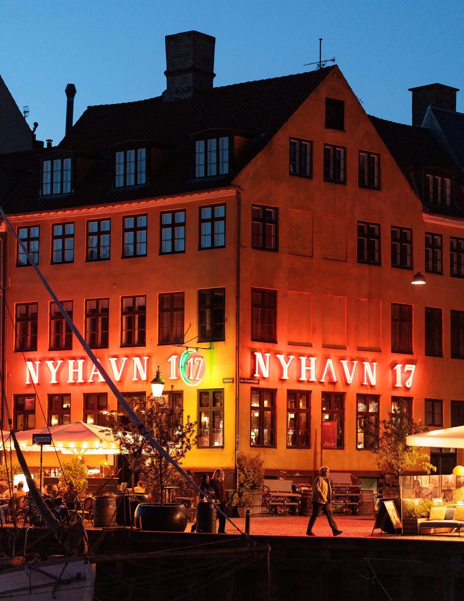 Night view of a building with illuminated 'NYHAVN 17' sign, outdoor seating, and people walking nearby, with trees and street lamps in the foreground.