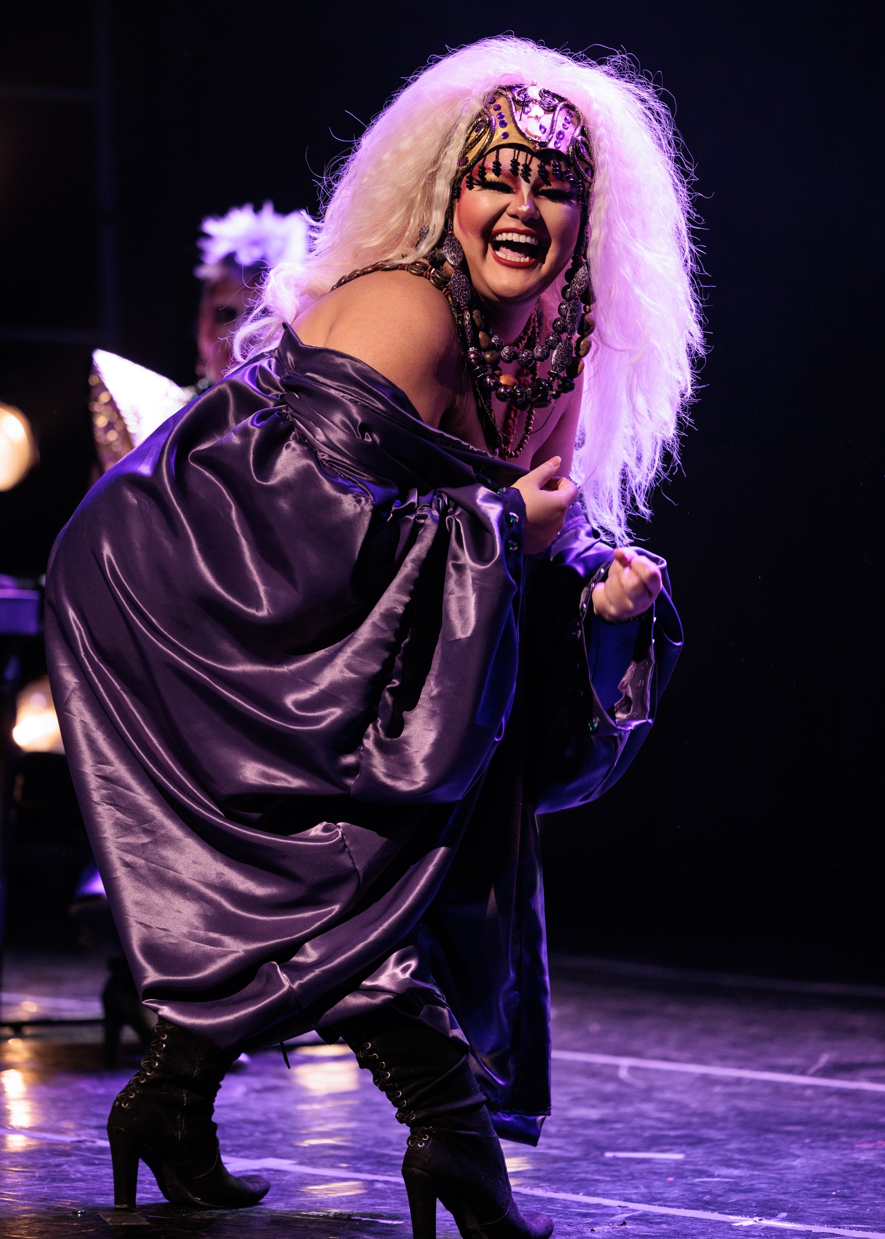 Performer with platinum blonde curly hair, wearing ornate headpiece, layered necklaces, black lace earrings, purple satin gown, and high-black boots, singing passionately on stage.