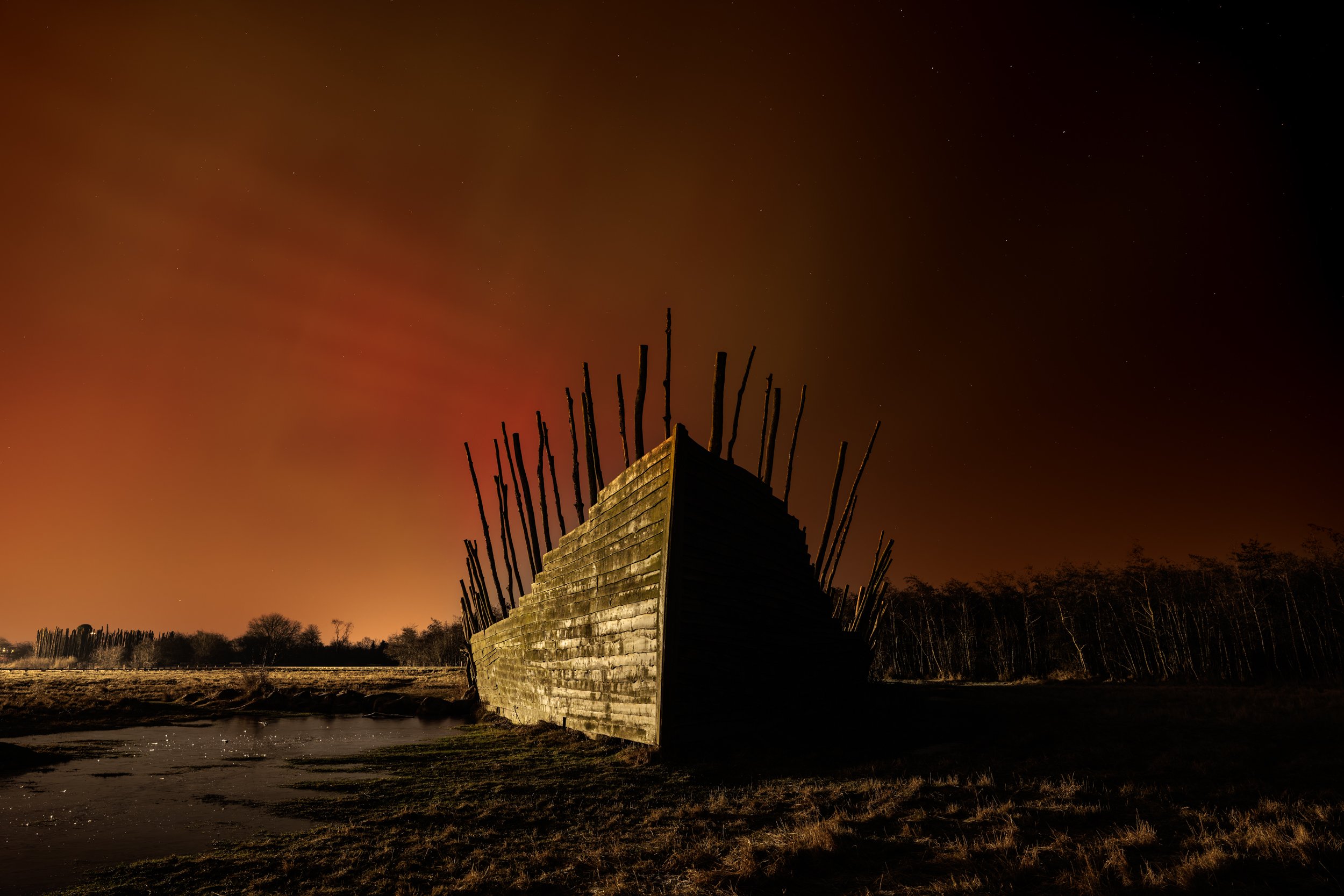 Nighttime view of a wooden structure with vertical sticks on top, set against a dark sky with stars, near a water body and trees in the background.
