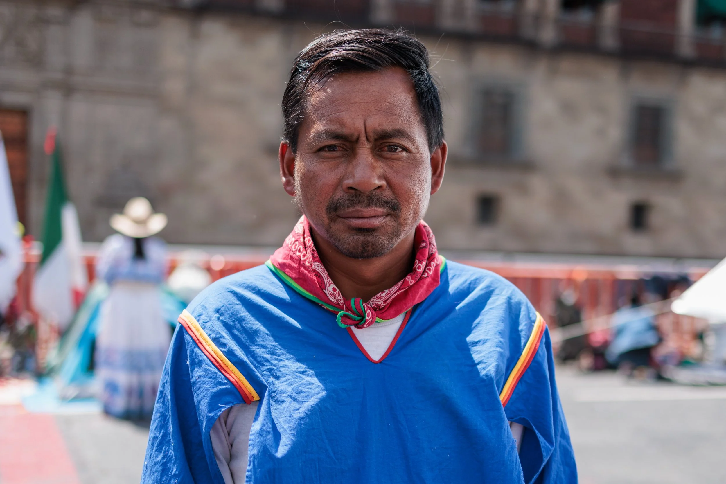 A man wearing a blue costume with rainbow trim and a red bandana around his neck, standing outdoors with a blurred background of buildings and people.
