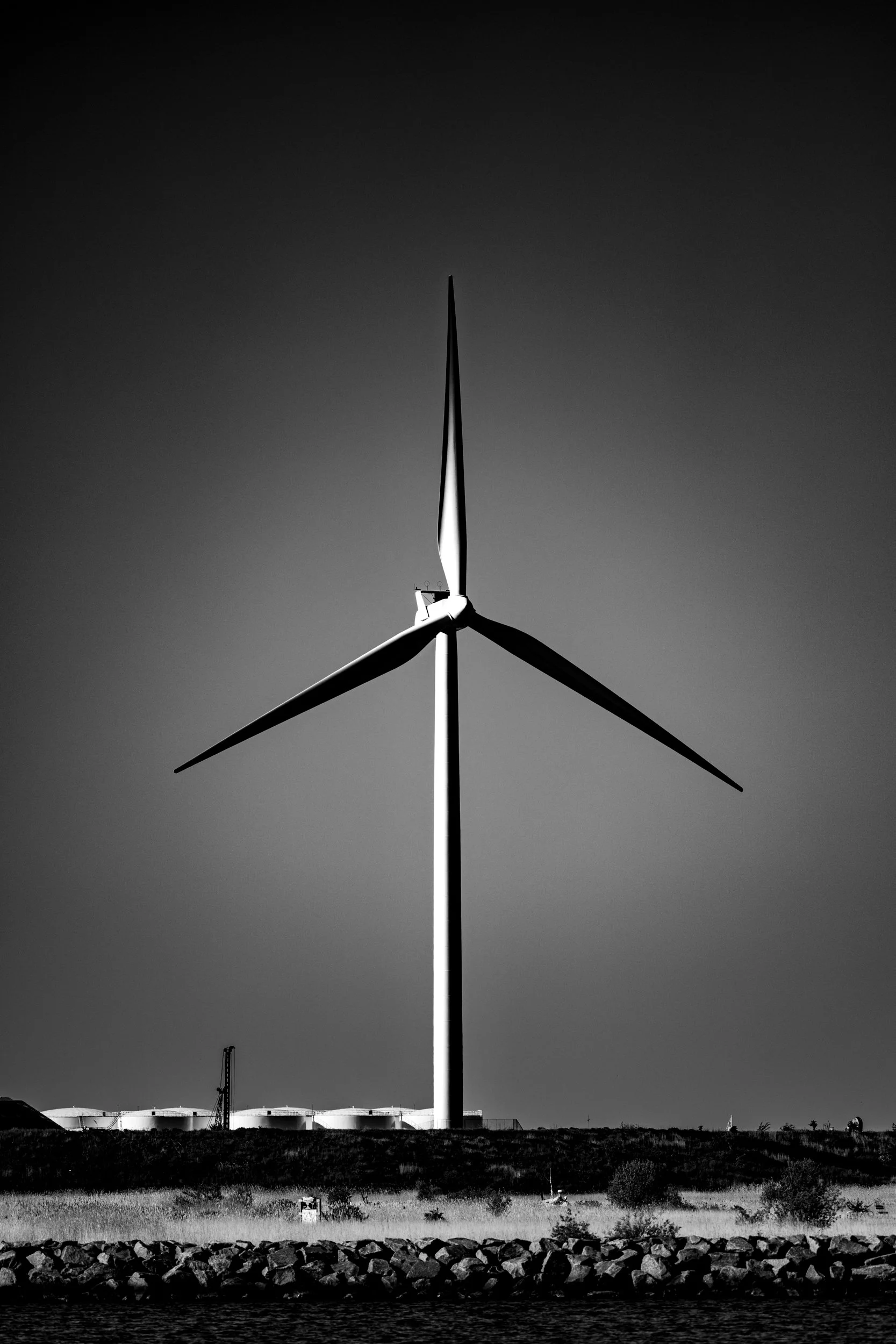 Black and white photo of a large wind turbine with three blades, standing on land against a clear sky, with industrial structures and trees in the background.