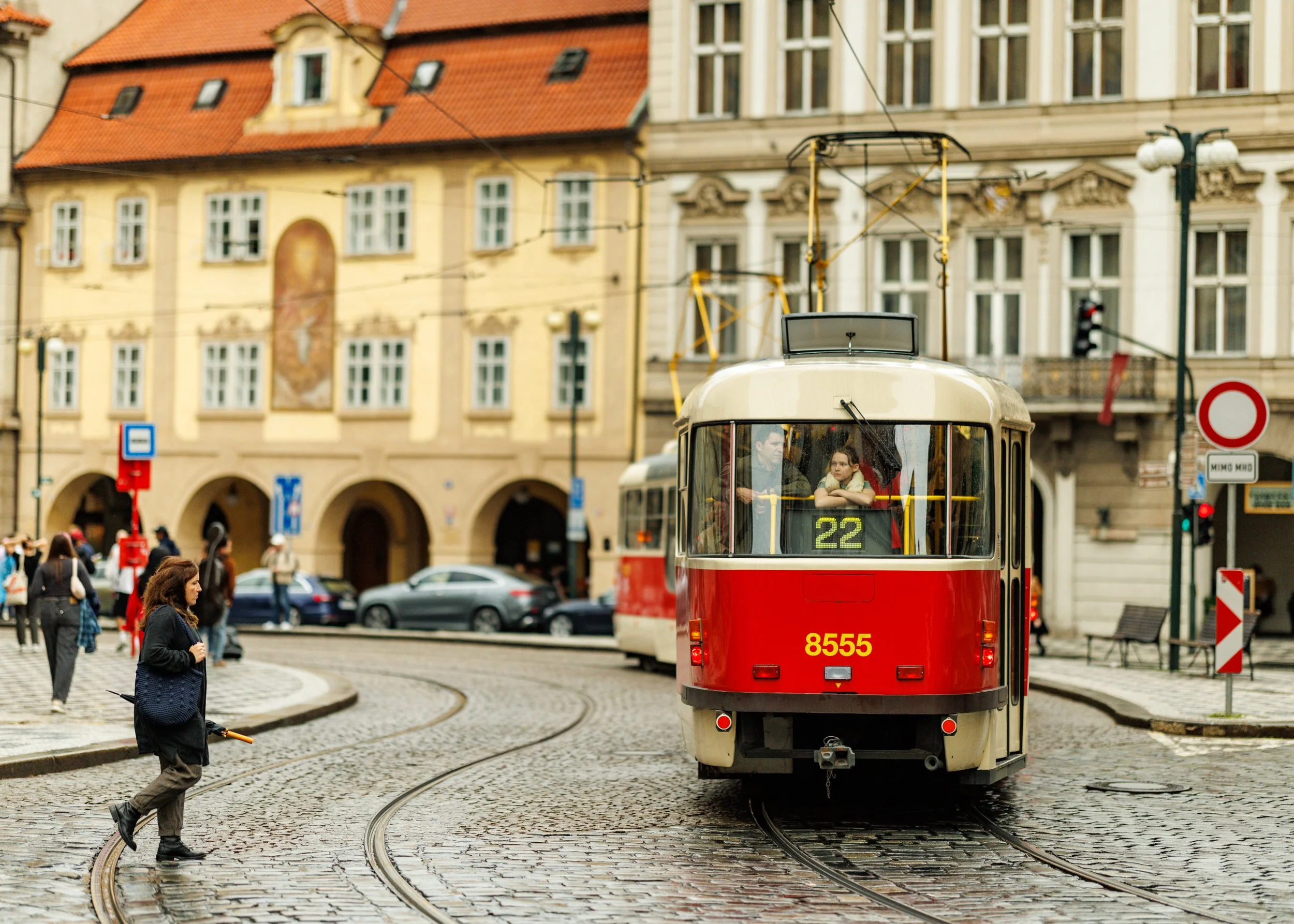 A red and white tram with the number 8555 on the back, moving along cobblestone tram tracks on a city street. Pedestrians are crossing the street, and buildings with ornate facades and red-tiled roofs are visible in the background.