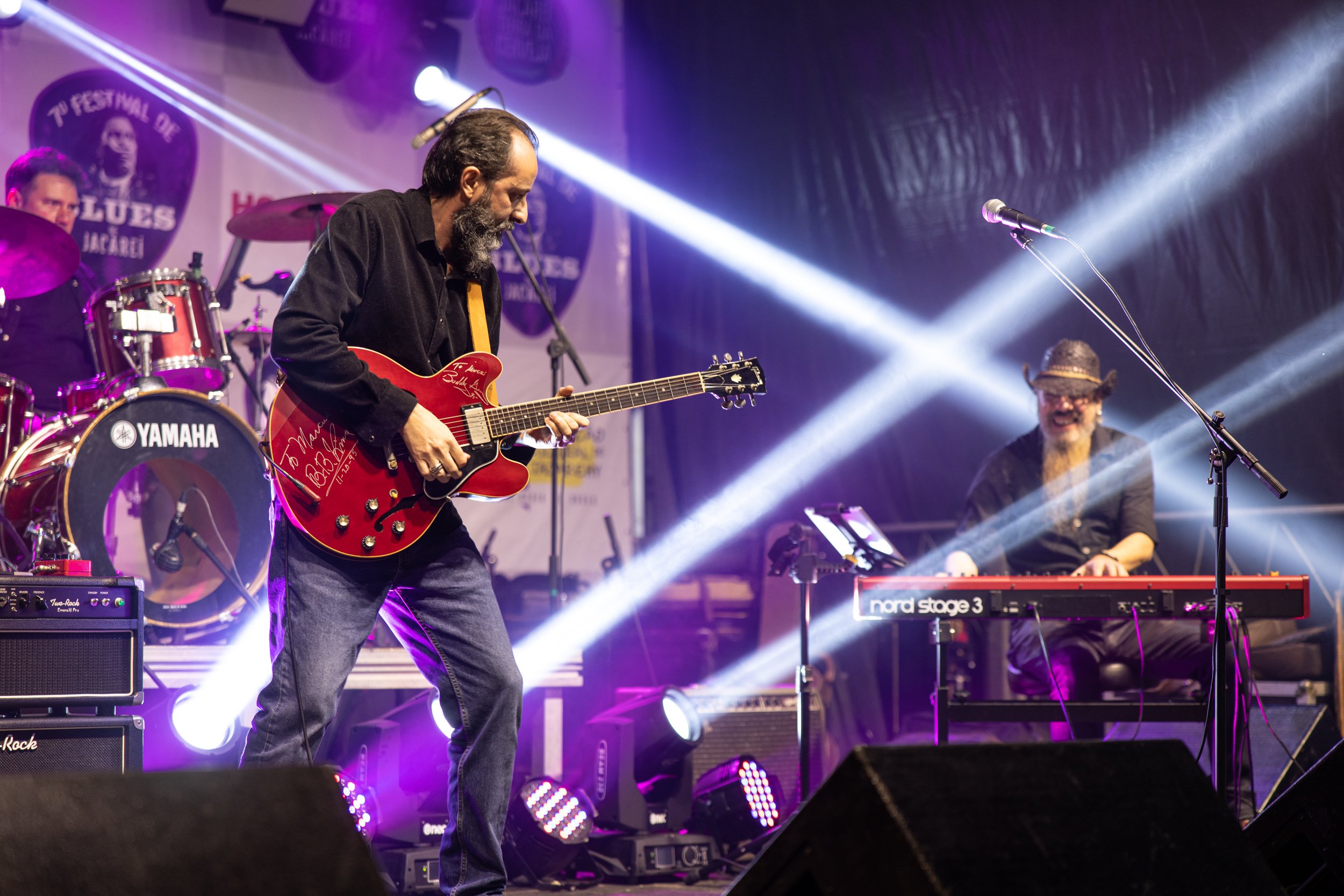 Musicians performing on stage during a concert with colorful stage lighting, including a guitarist, a drummer in the background, and a keyboard player on the right.