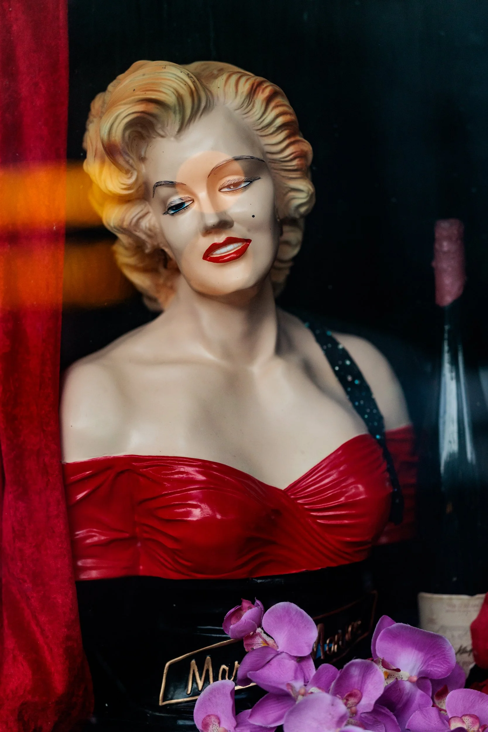 A Marilyn Monroe mannequin with wavy blonde hair, red lipstick, and a strapless red dress, surrounded by purple orchids and a red curtain