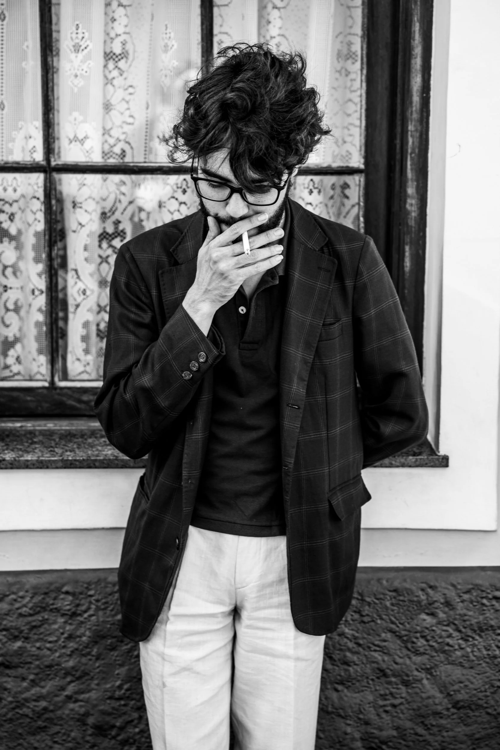 A man with curly hair and glasses, wearing a dark blazer over a dark shirt, standing outdoors in front of a window with lace curtains, holding a lit cigarette near his mouth, looking down.