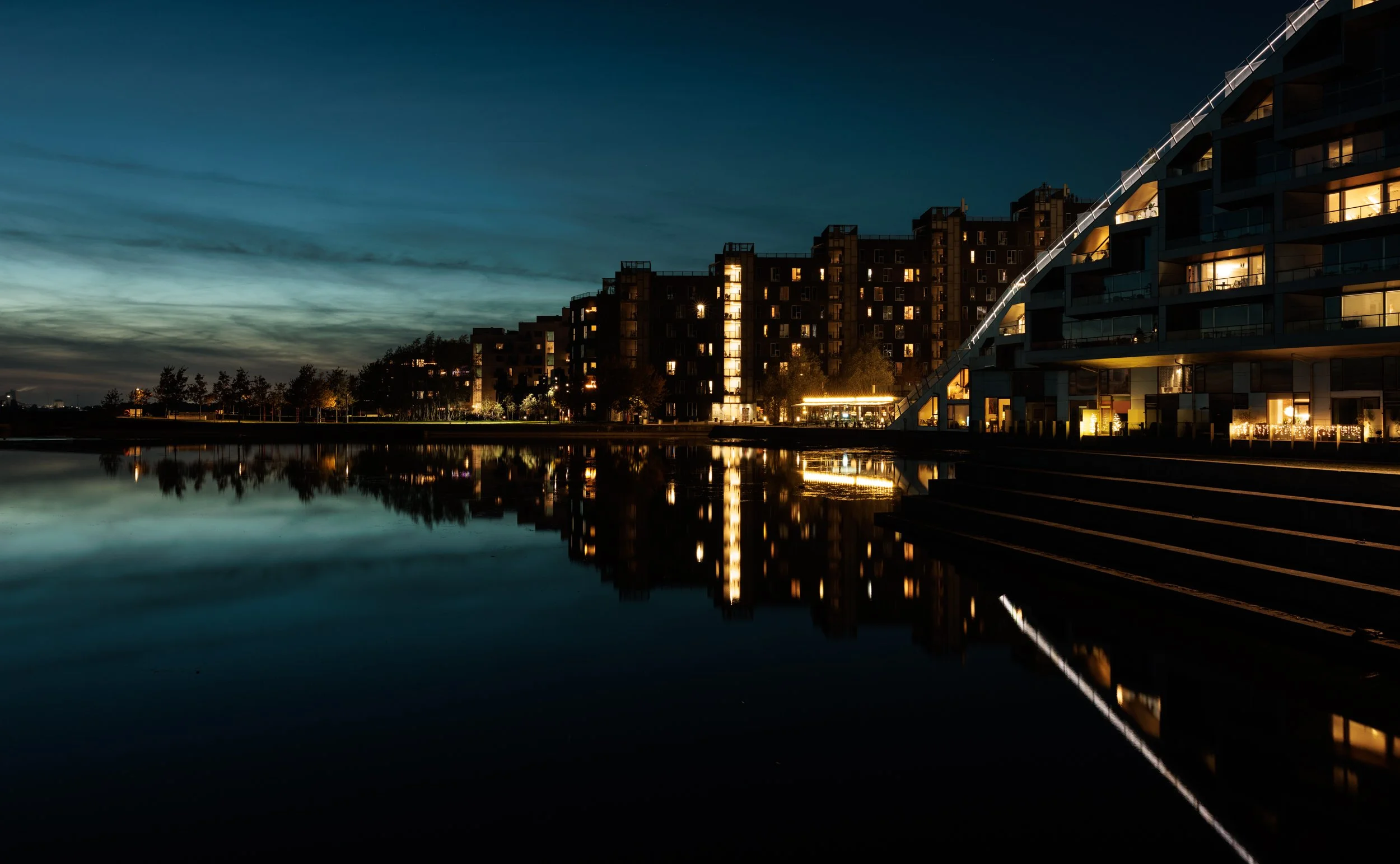 Night view of modern apartment buildings with illuminated windows, reflecting on a calm body of water.