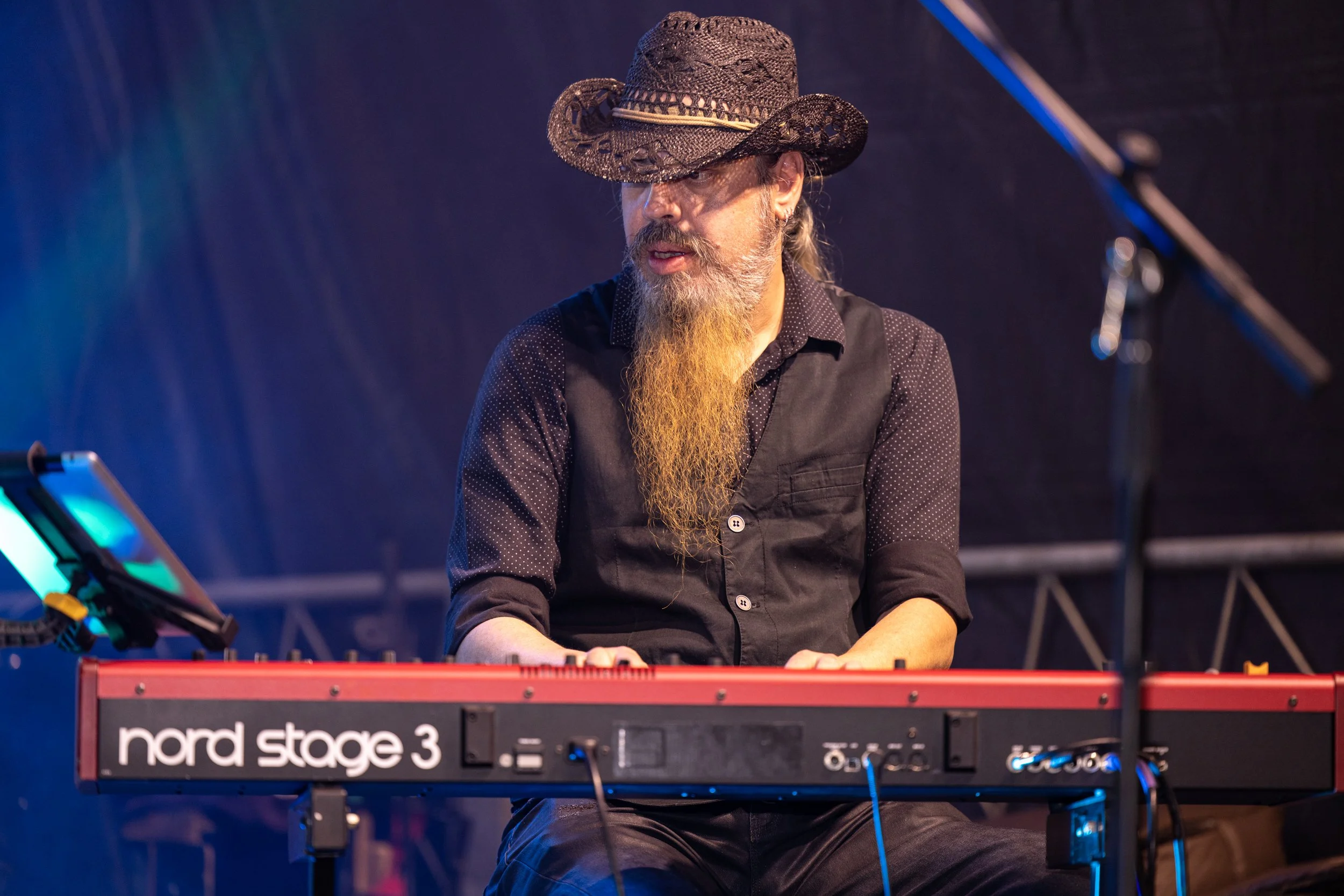 A man with a long beard wearing a black shirt and a cowboy hat plays a red Nord Stage 3 keyboard on stage.