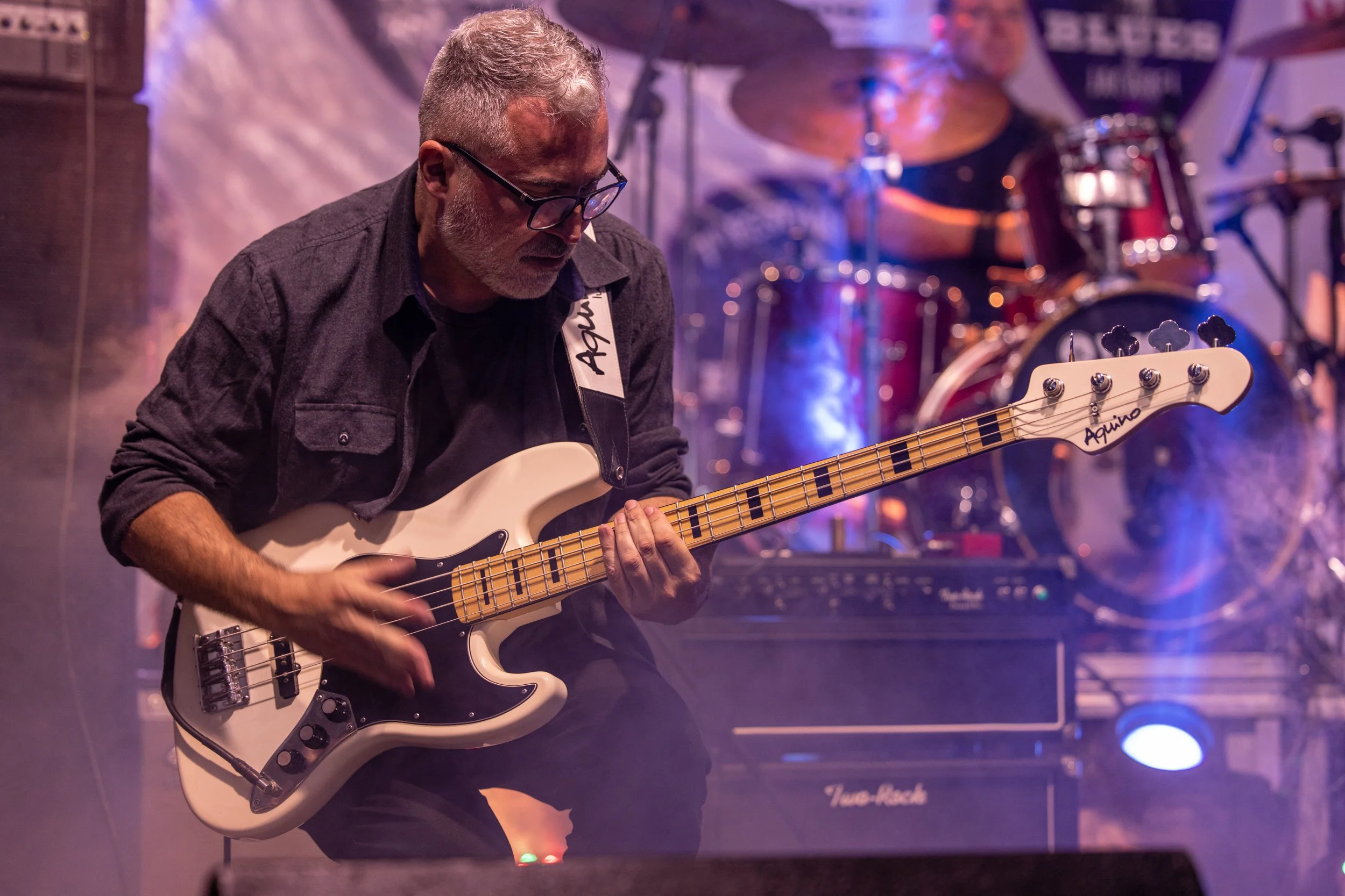 A middle-aged man with gray hair, glasses, and a beard playing an electric bass guitar on stage. He is wearing a dark shirt and appears focused. There are drums and an amplifier in the background with colorful stage lighting.