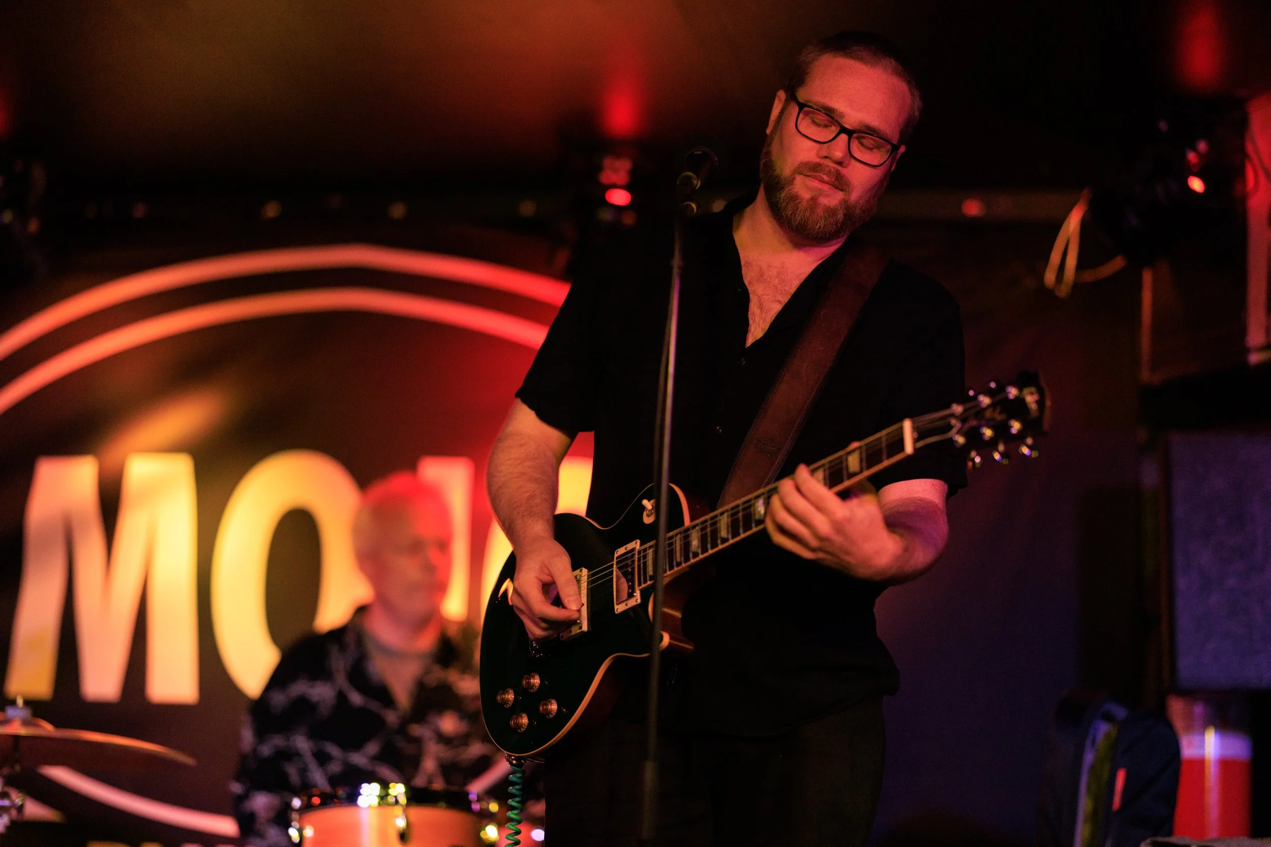 Man with glasses and beard playing an electric guitar at a dimly lit venue, neon lights and a drum set visible in the background.