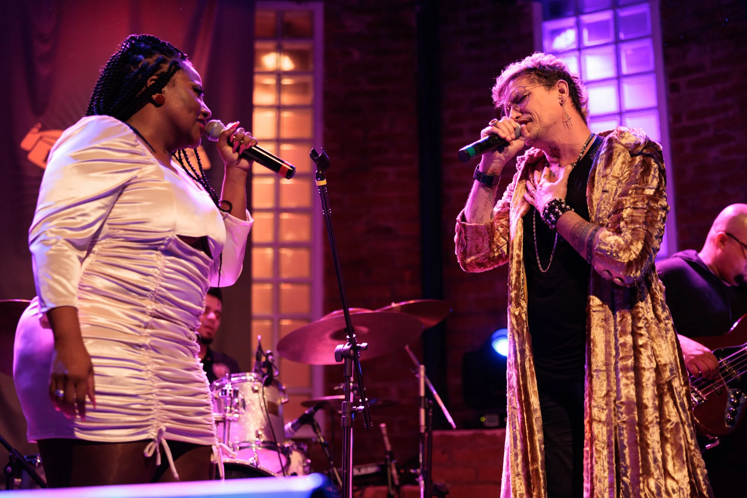 Two singers perform on stage, one woman with braids and white dress, and one man with glasses and patterned coat, with musical instruments and a brick wall background.