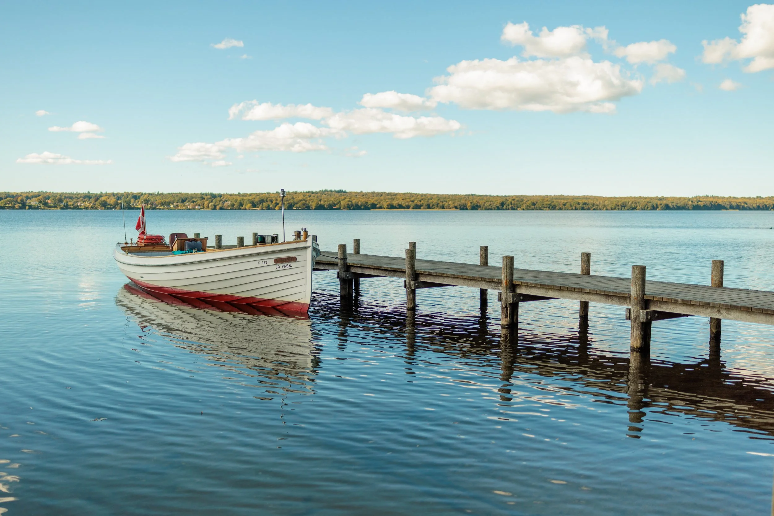 A boat docked at a wooden pier on a calm lake with clear blue sky and fluffy clouds, distant forested shoreline.
